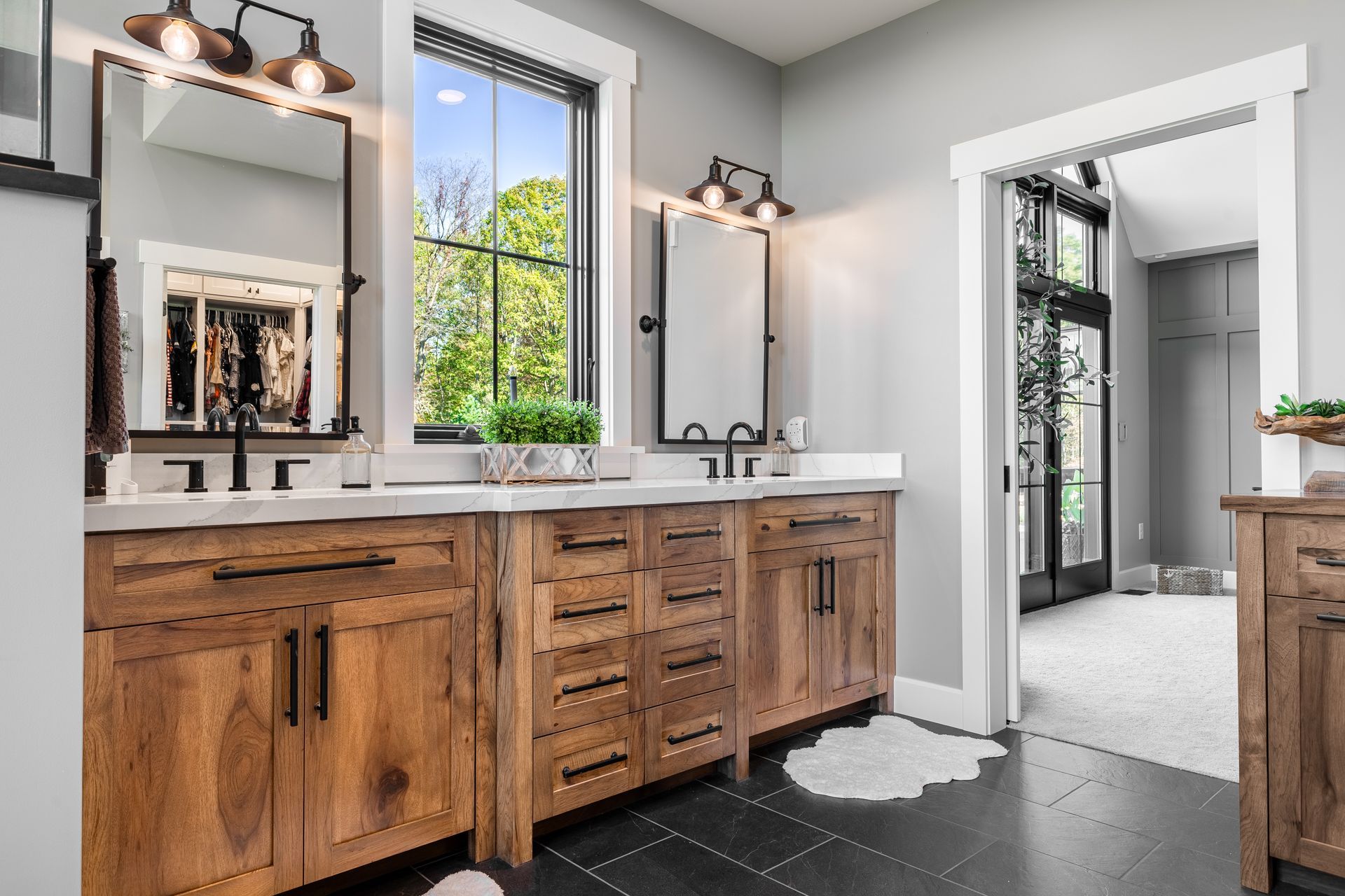 Bathroom with wood vanity, black fixtures, mirrors, and a window overlooking greenery.