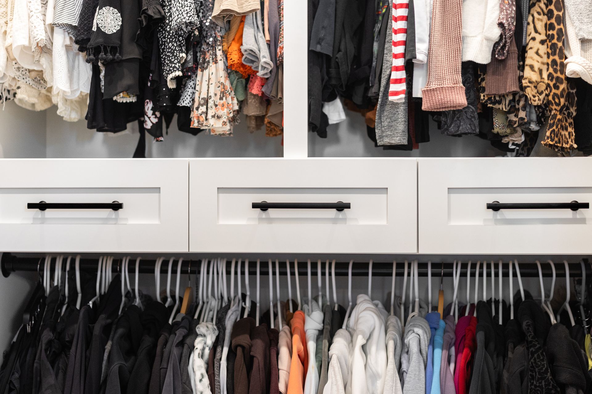 Closet with hanging clothes in various colors, above drawers with black handles.