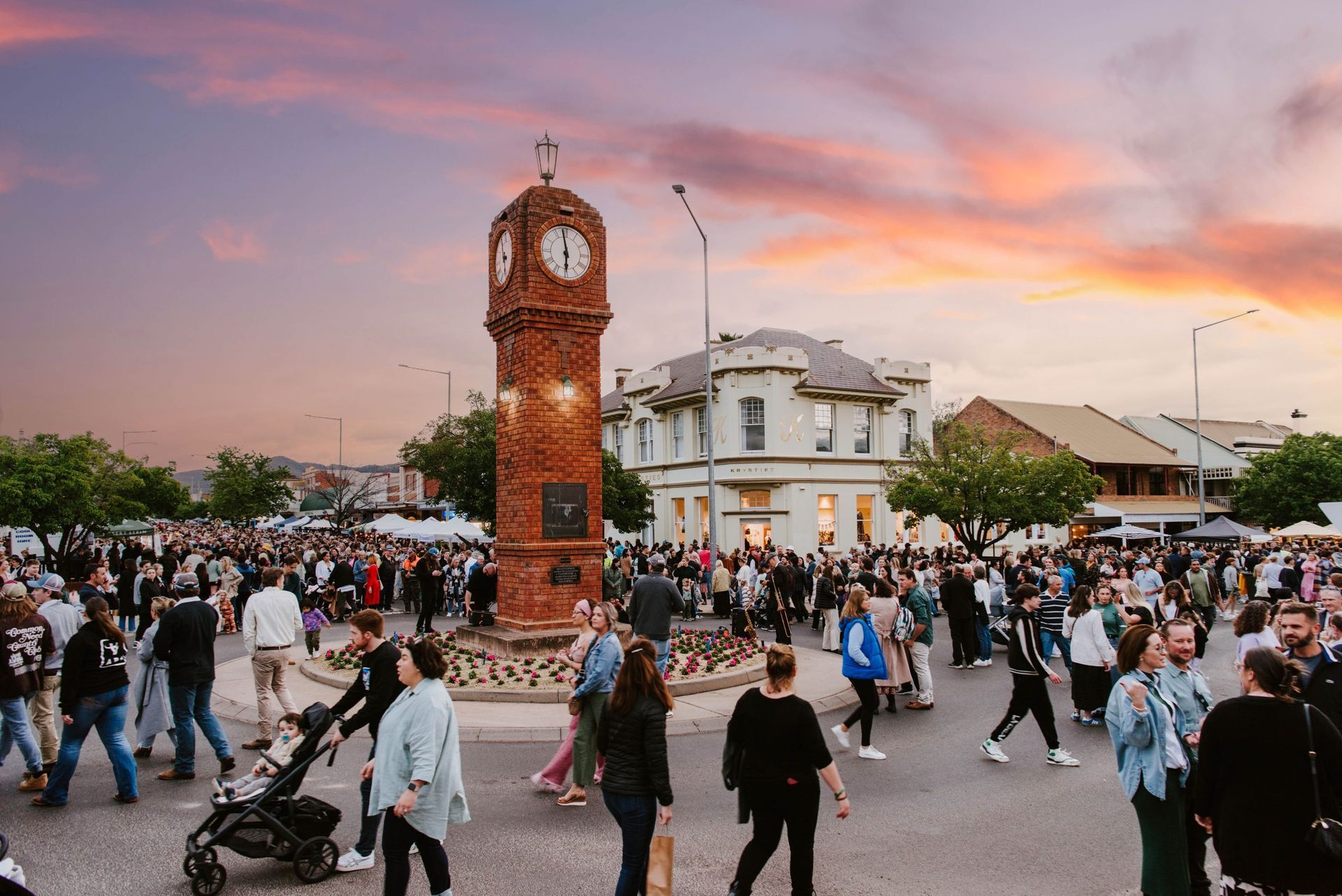 Crowd gathered around a brick clock tower in a town square at sunset— Mudgee Passenger Transport in Mudgee, NSW