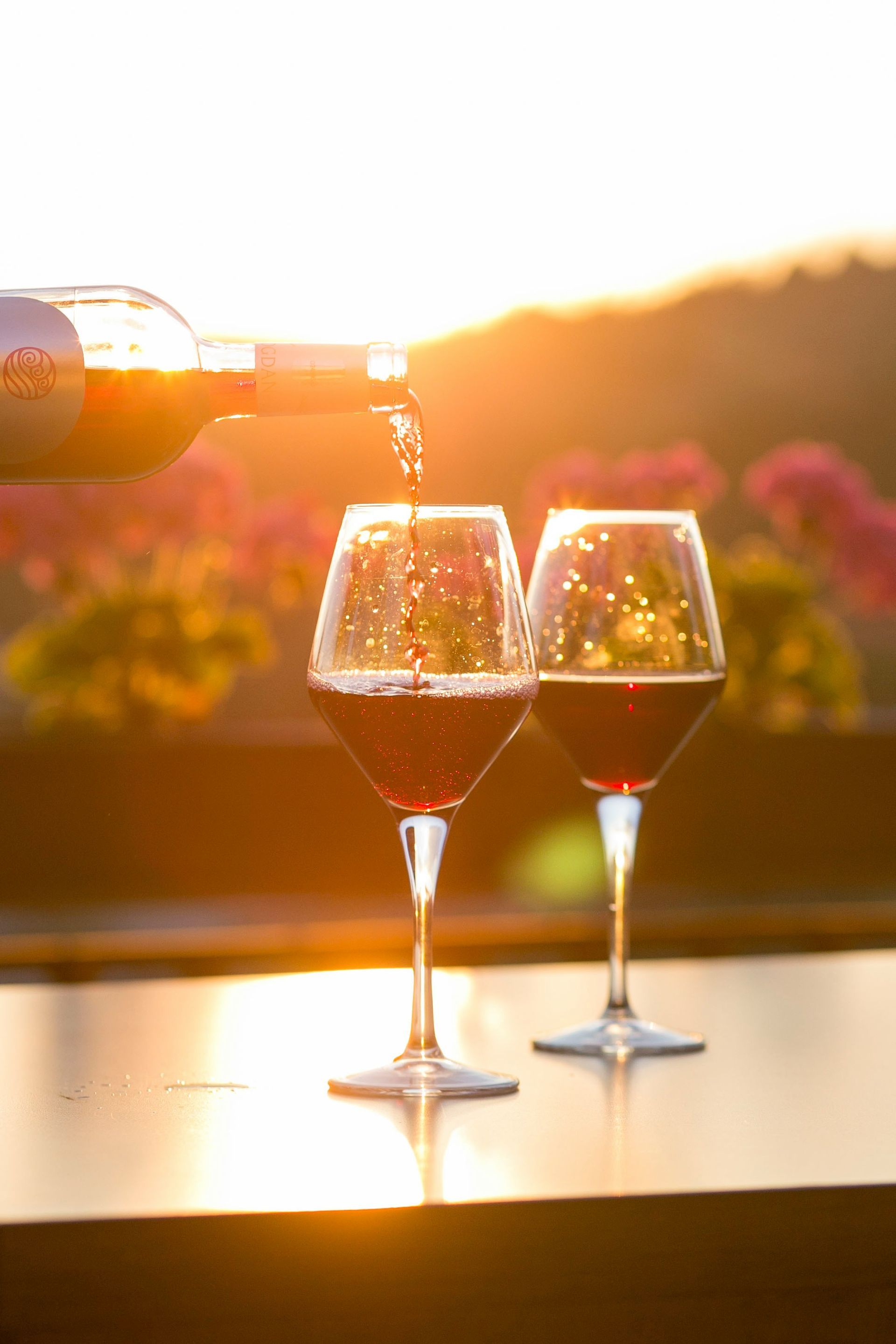 Two wine glasses on a sunlit table with flowers and a warm sunset in the background