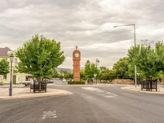 Quiet street with a red brick clock tower at a roundabout, flanked by trees and low buildings under cloudy skies