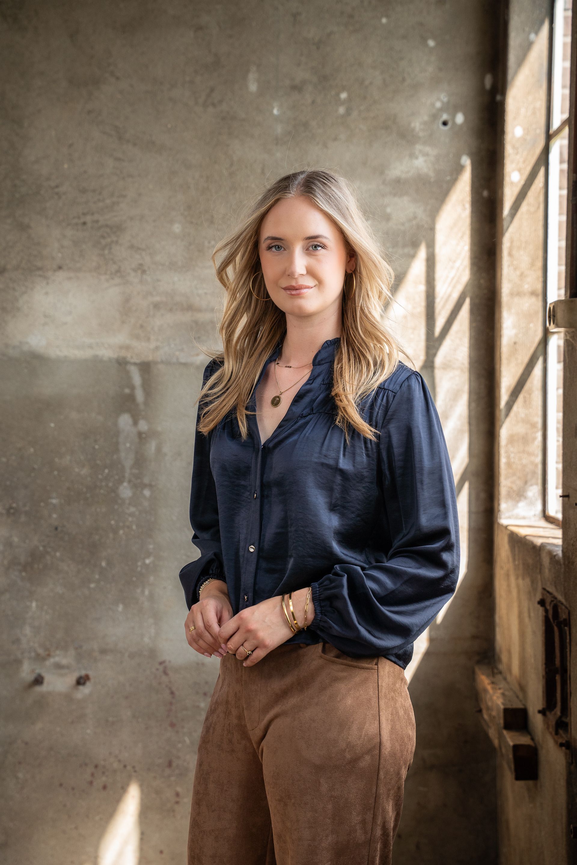Vrouw staat bij een raam in een rustieke kamer, gekleed in een donkerblauwe blouse en een bruine broek.