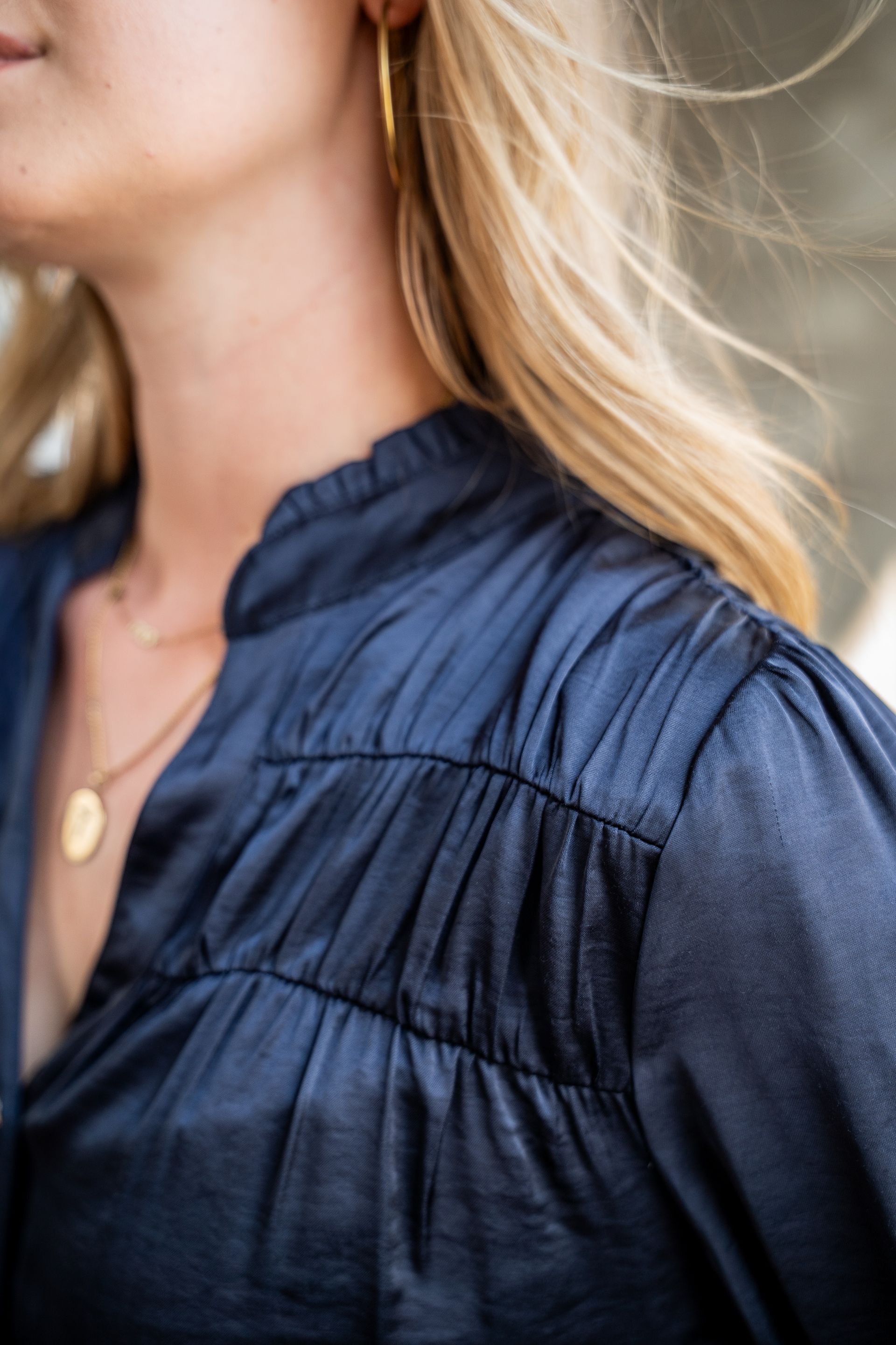 Vrouw in een donkerblauwe blouse met geplooide mouwen en een gouden hangerketting, van dichtbij gefotografeerd.