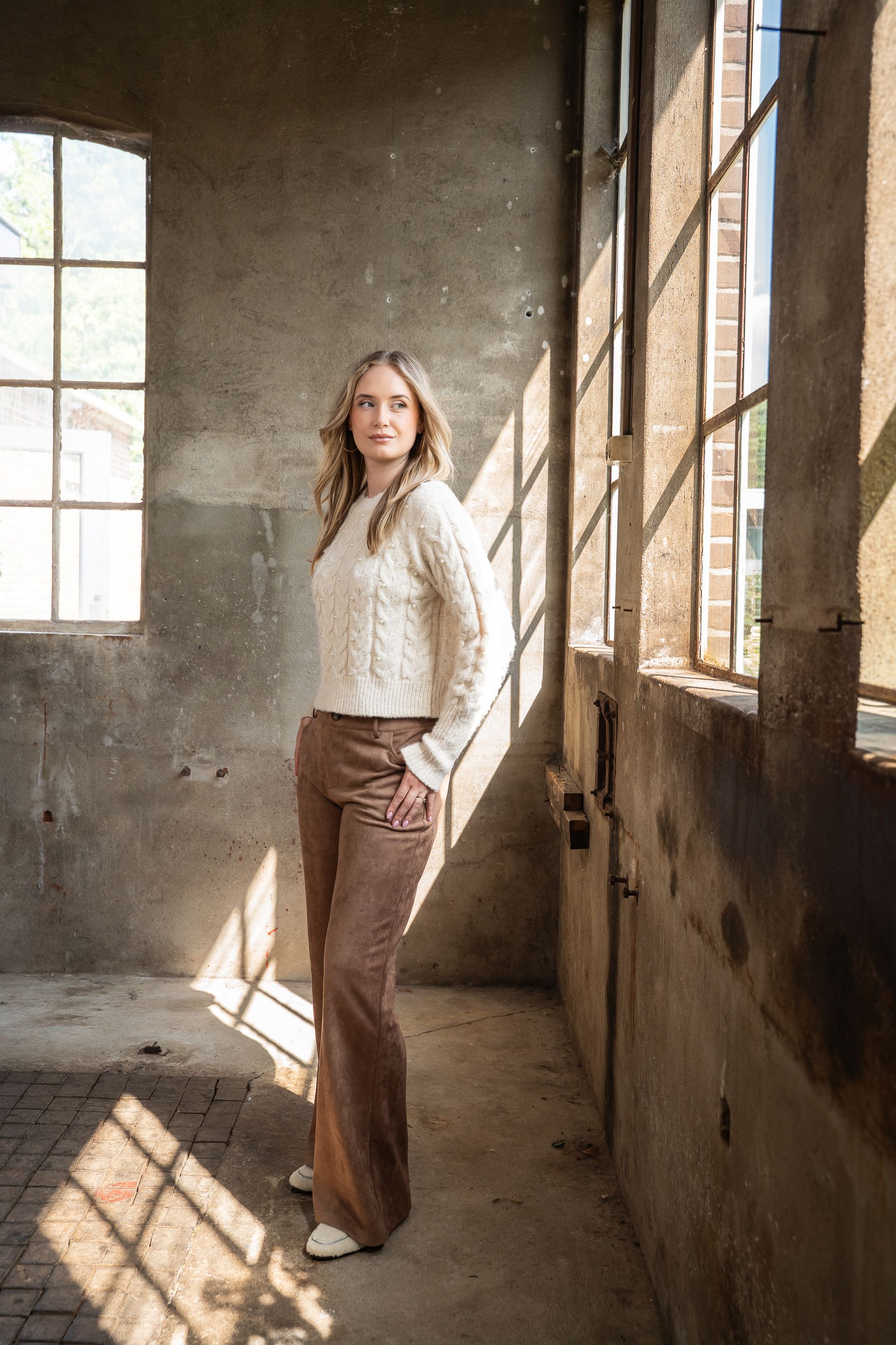 Vrouw in een witte trui en bruine broek staat in een zonovergoten industriële ruimte bij grote ramen.