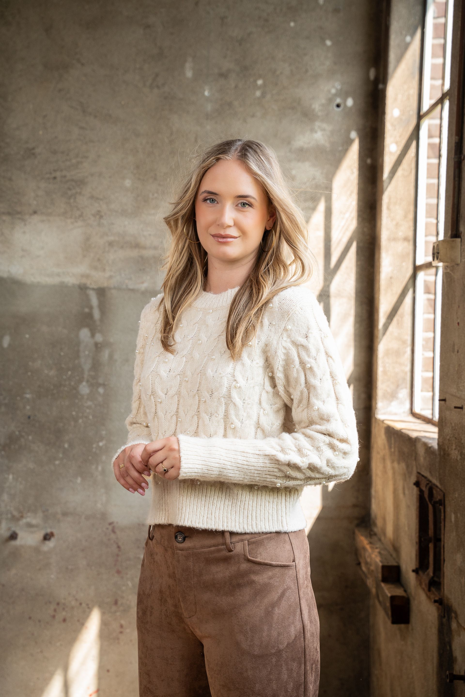 Vrouw in een crèmekleurige trui en bruine broek staat bij een raam in een rustieke kamer.