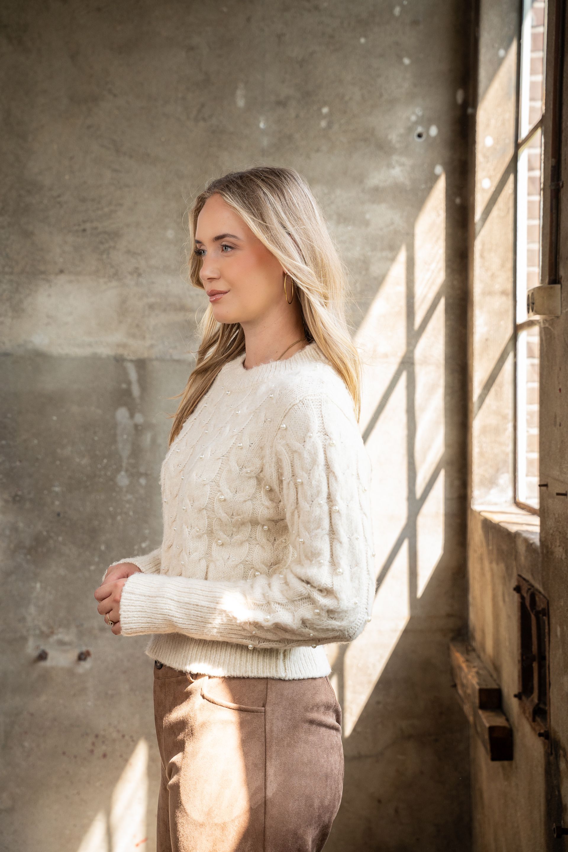 Vrouw in een witte trui staat bij een zonnig raam in een rustieke kamer.
