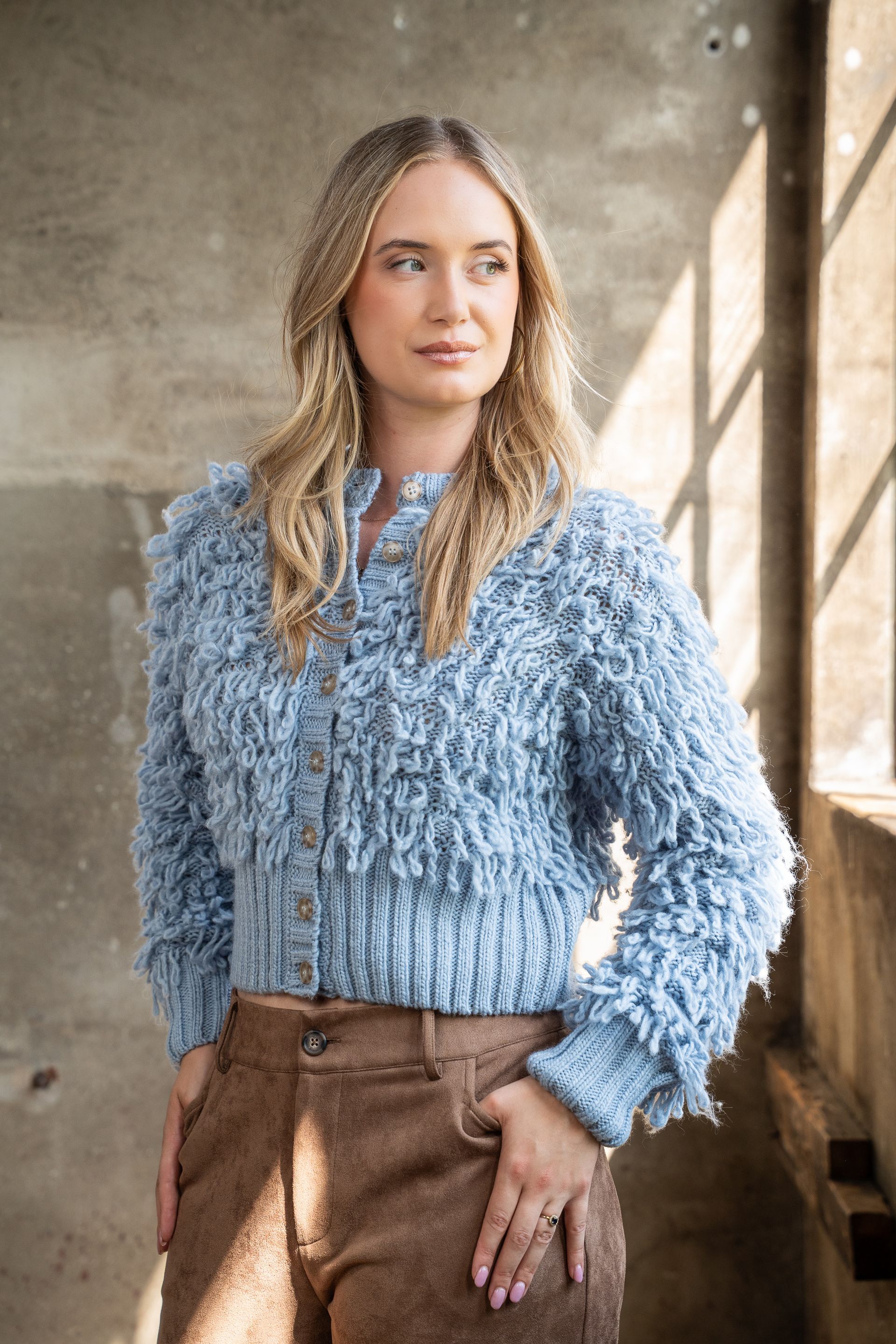 Vrouw in een blauwe, gestructureerde trui en een bruine broek staat bij een raam in het zachte licht.