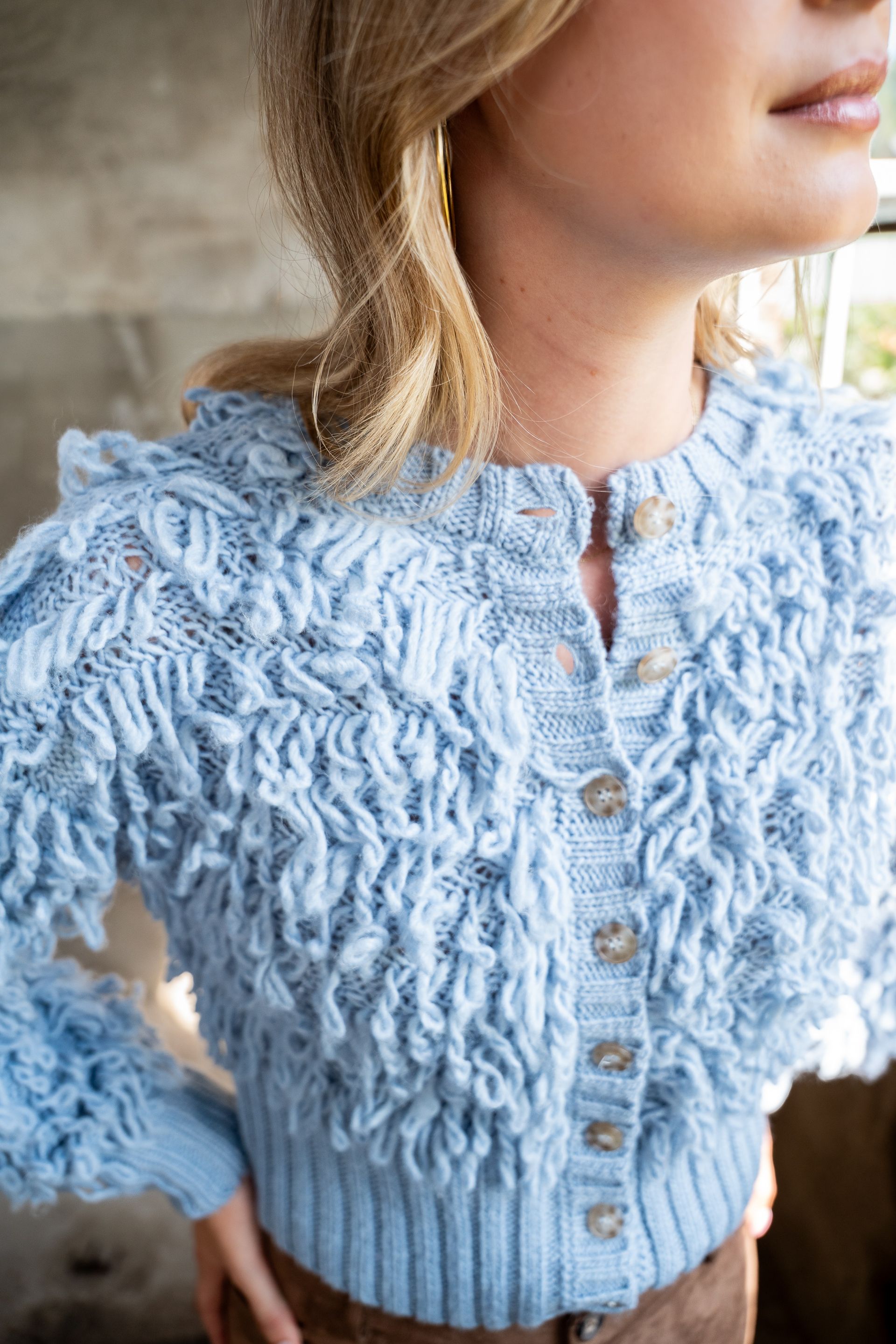 Lichtblauwe, gestructureerde, met knoopjes versierde cardigan met ruches, gedragen door een persoon binnenshuis.