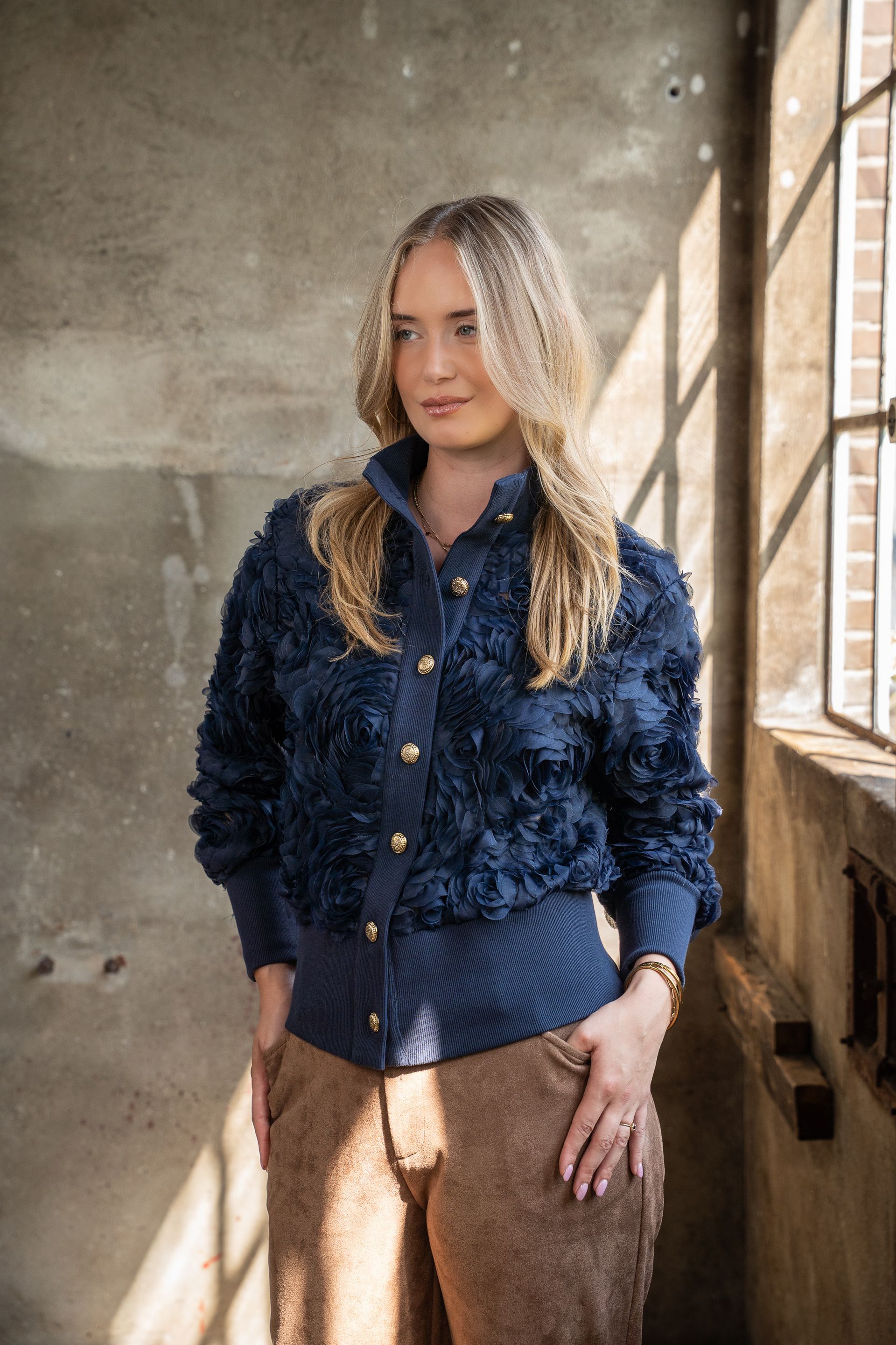Vrouw in een donkerblauwe jas met textuur en een blauwe blouse staat bij een raam in een zonnige kamer.