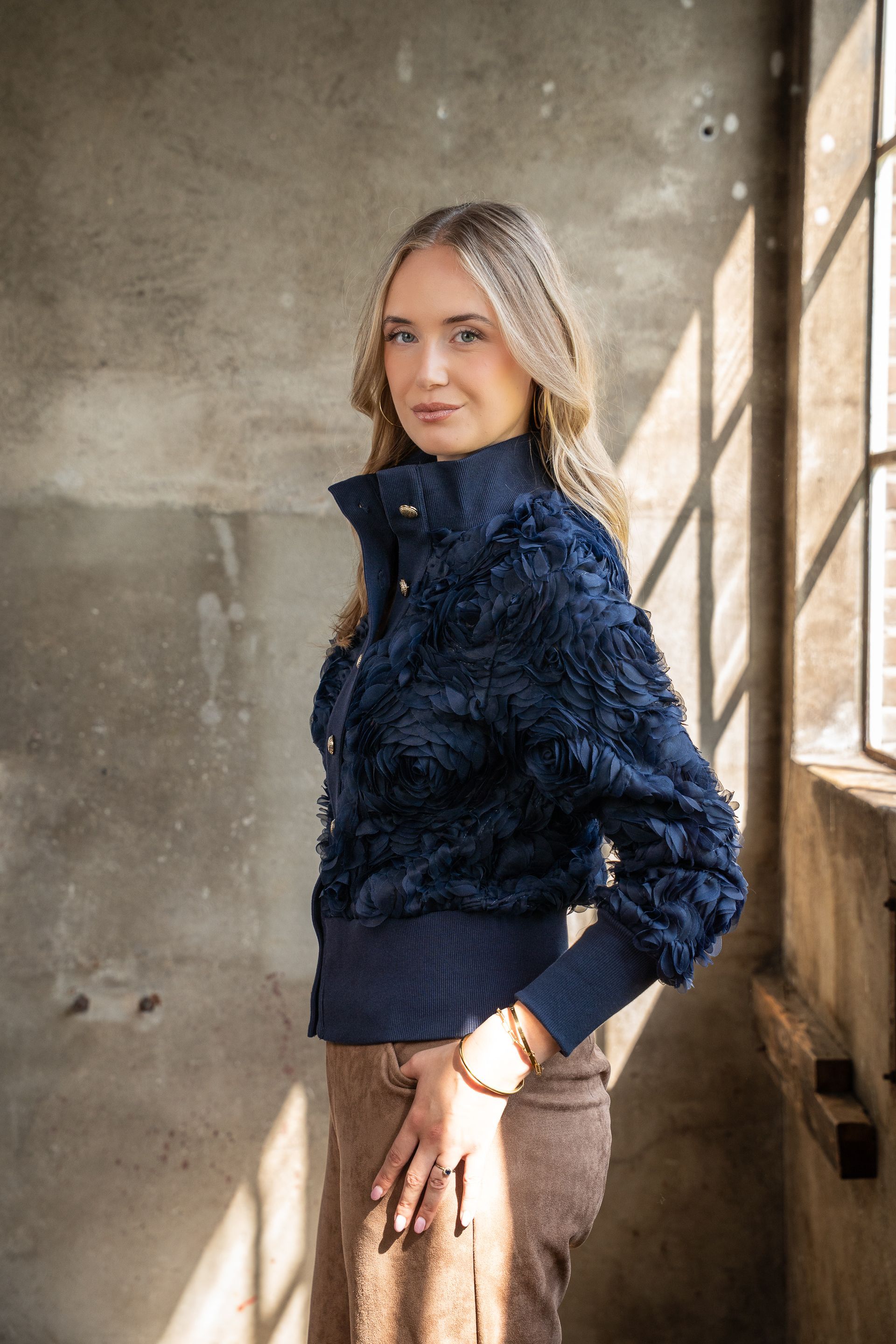 Vrouw in een donkerblauwe jas met textuur poseert bij een zonnig raam in een rustieke kamer.