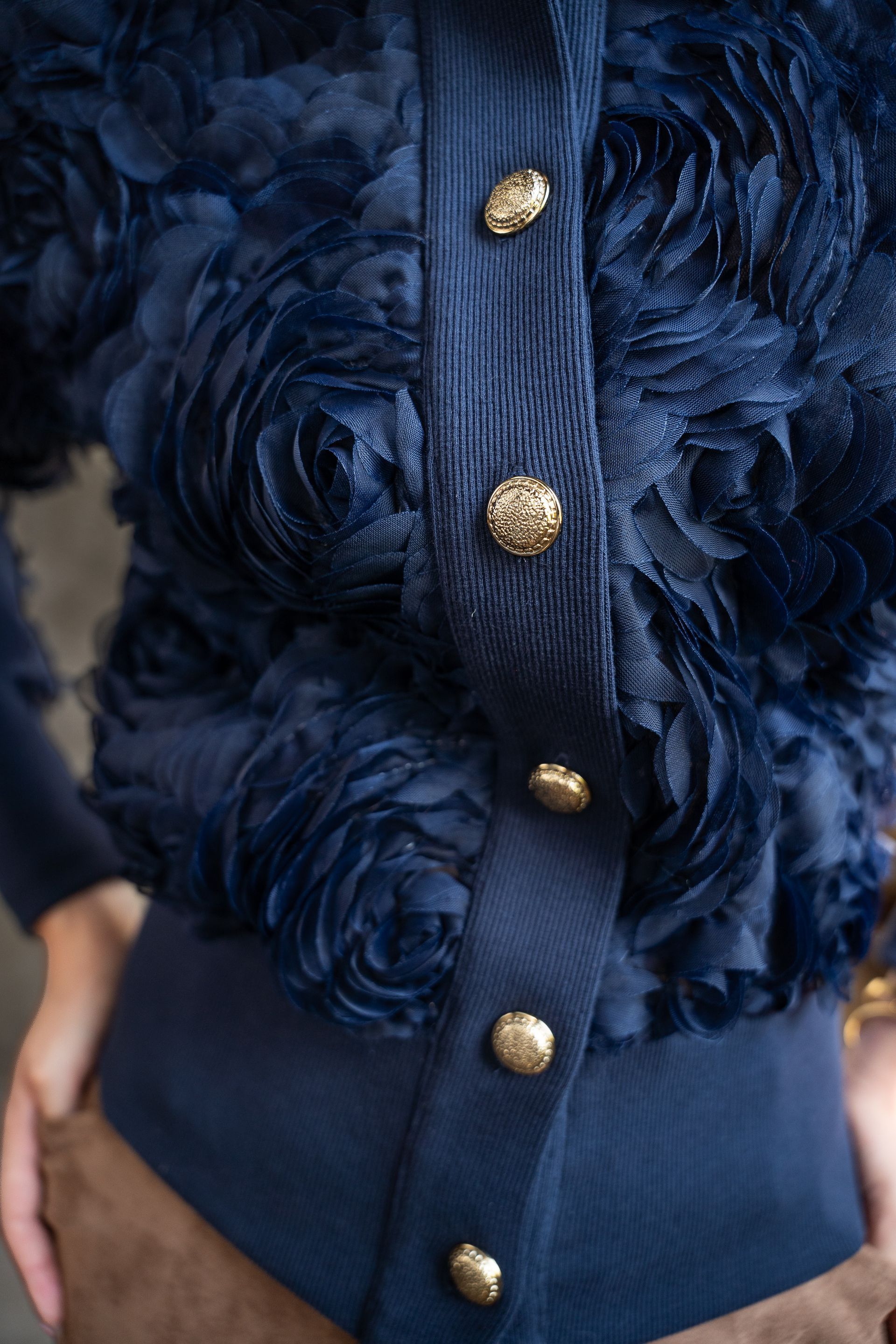 Detailopname van een marineblauwe jas met textuur, gouden knopen en een geribbelde taille.