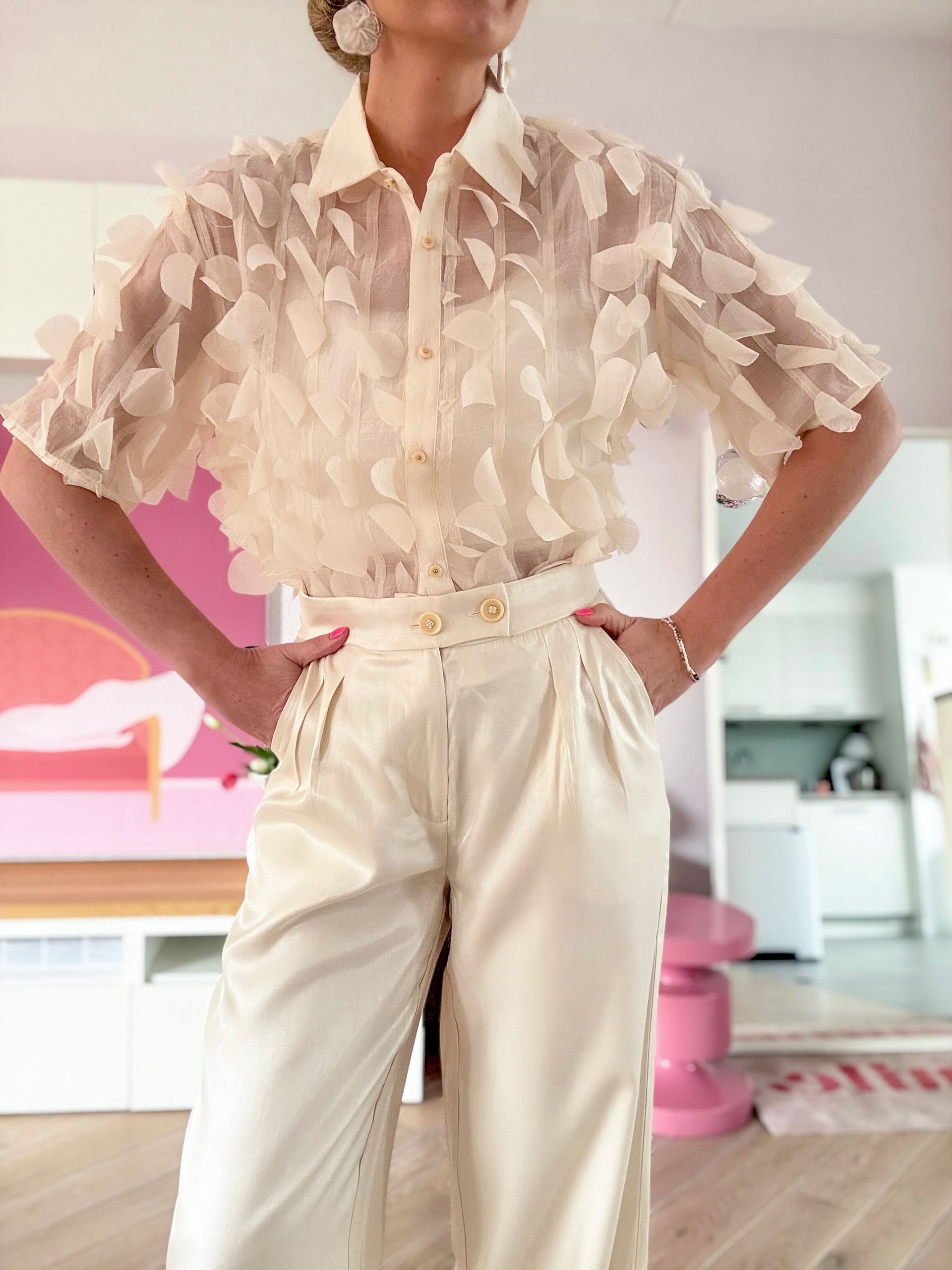 Een persoon in een crèmekleurige blouse met ruches en een satijnen broek staat met de handen in de zij in een kamer met roze accenten.