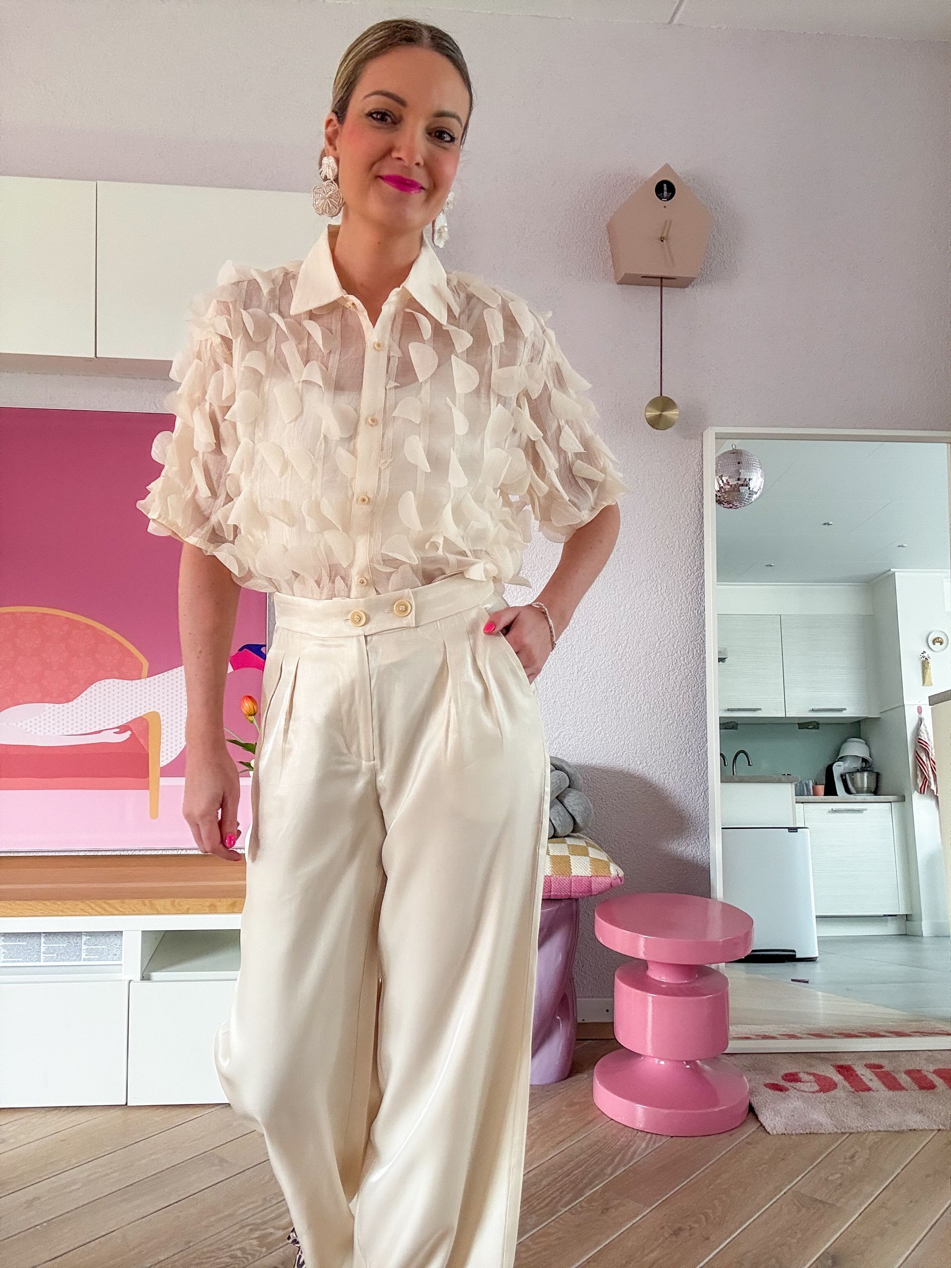 Vrouw staat binnen in een witte blouse met ruches en een crèmekleurige broek, poserend in een roze-witte kamer.