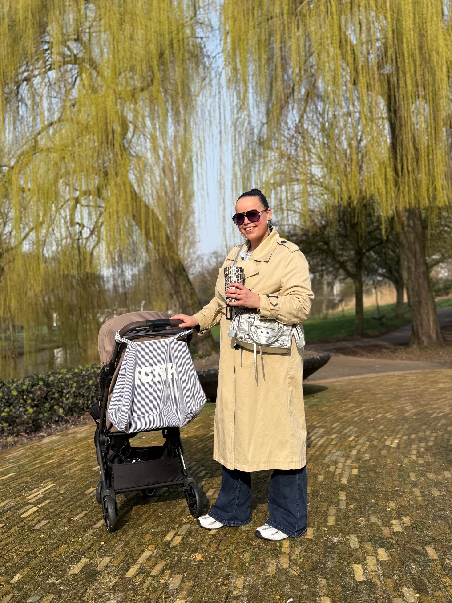 Een persoon in een beige jas staat naast een kinderwagen onder wilgen in een park.