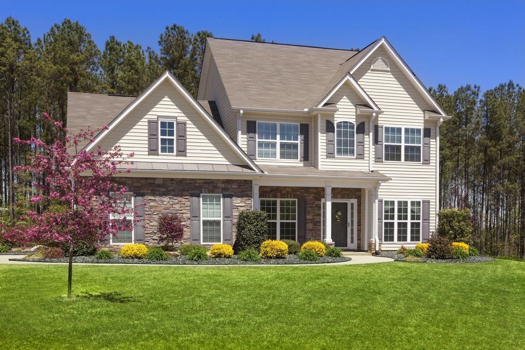 Uprooted tree lying on a grassy lawn. Two-story beige house with white trim, front porch, and blooming shrubs beside a green lawn.