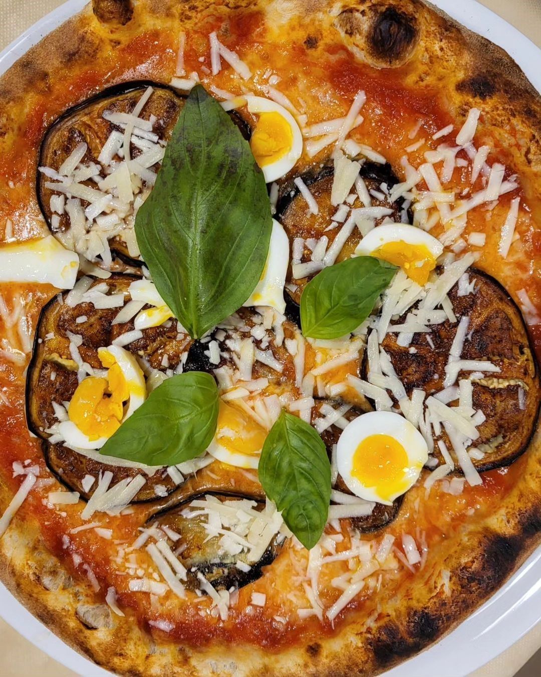 Primo piano di una pizza condita con basilico, pomodori a fette, uova e formaggio grattugiato su un piatto bianco.