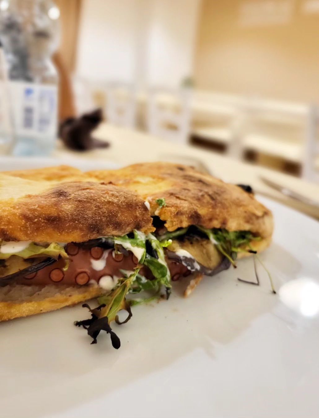 Un panino su un piatto bianco in un caffè luminoso, con verdure a foglia verde e ripieno ben visibile.