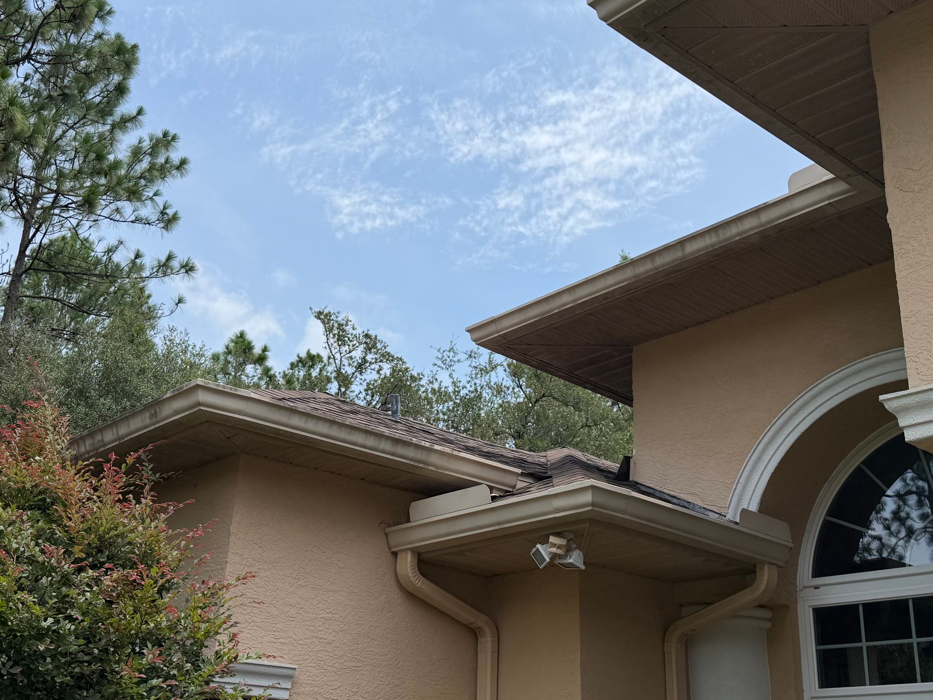 Suburban house roofline with beige stucco walls, arched entry, and trees against a blue sky
