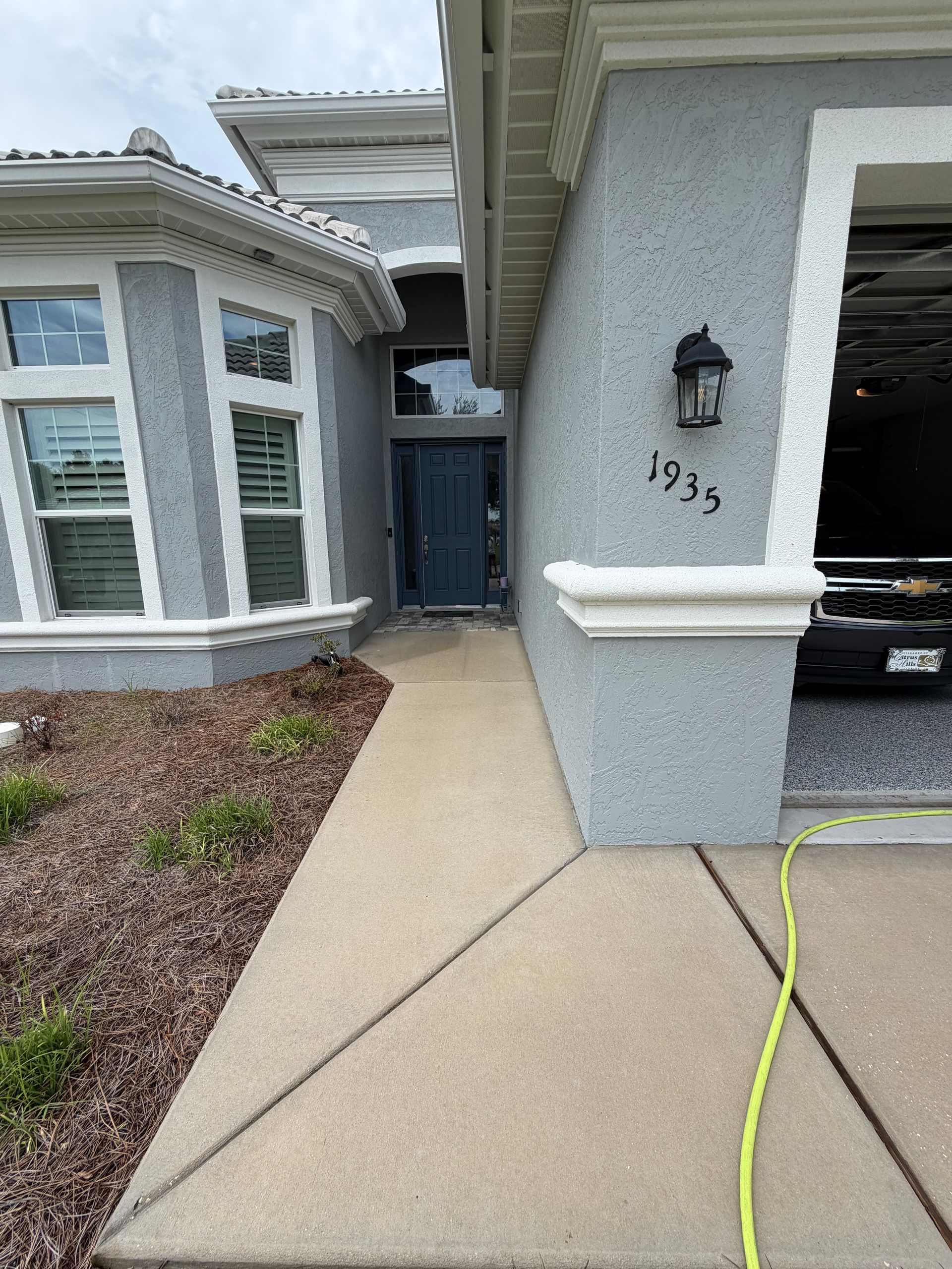 Front entrance of a house with gray stucco walls, bay windows, driveway, and house number 1995.