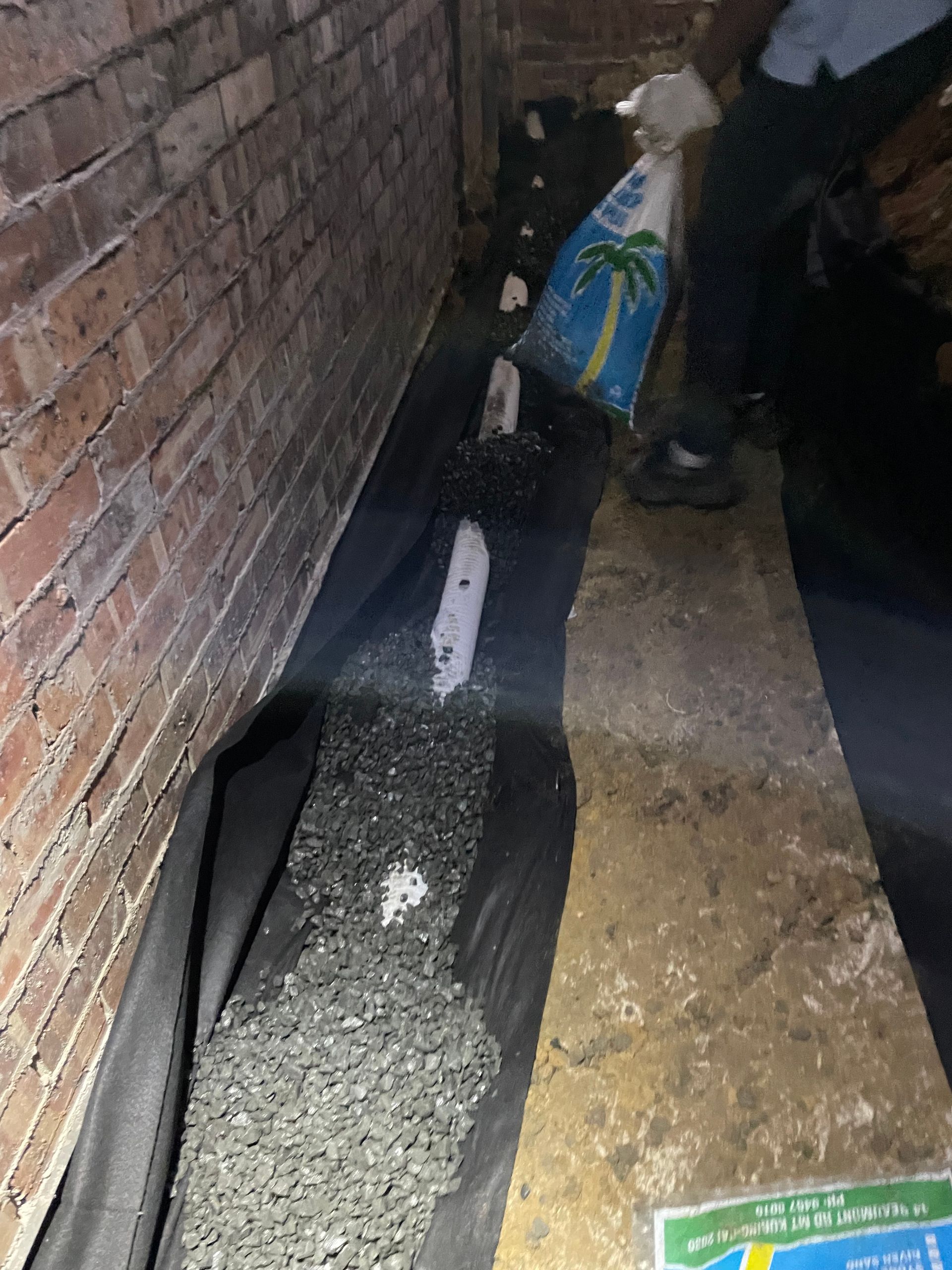 Person pouring gravel from a bag onto a black ground cover along a brick wall in a basement.