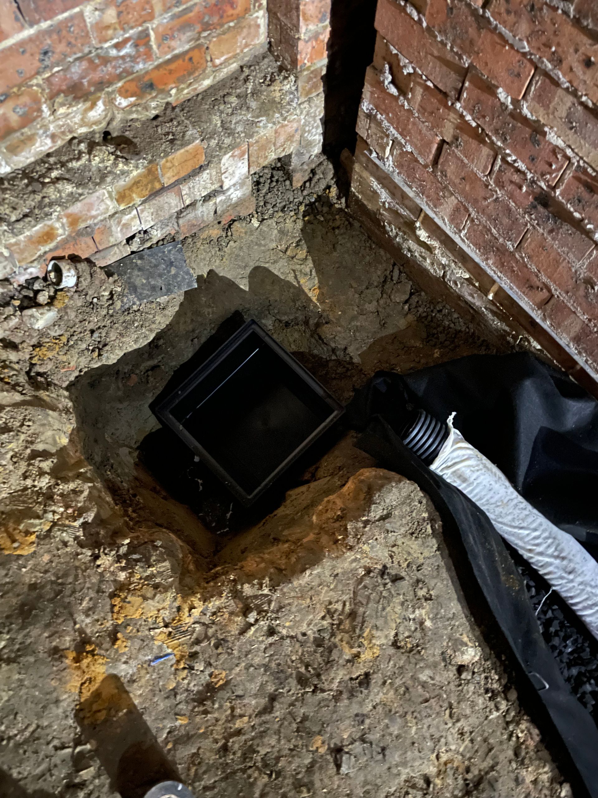 Excavated area near brick walls with a black basin and drainage pipe. Earth tones dominate.
