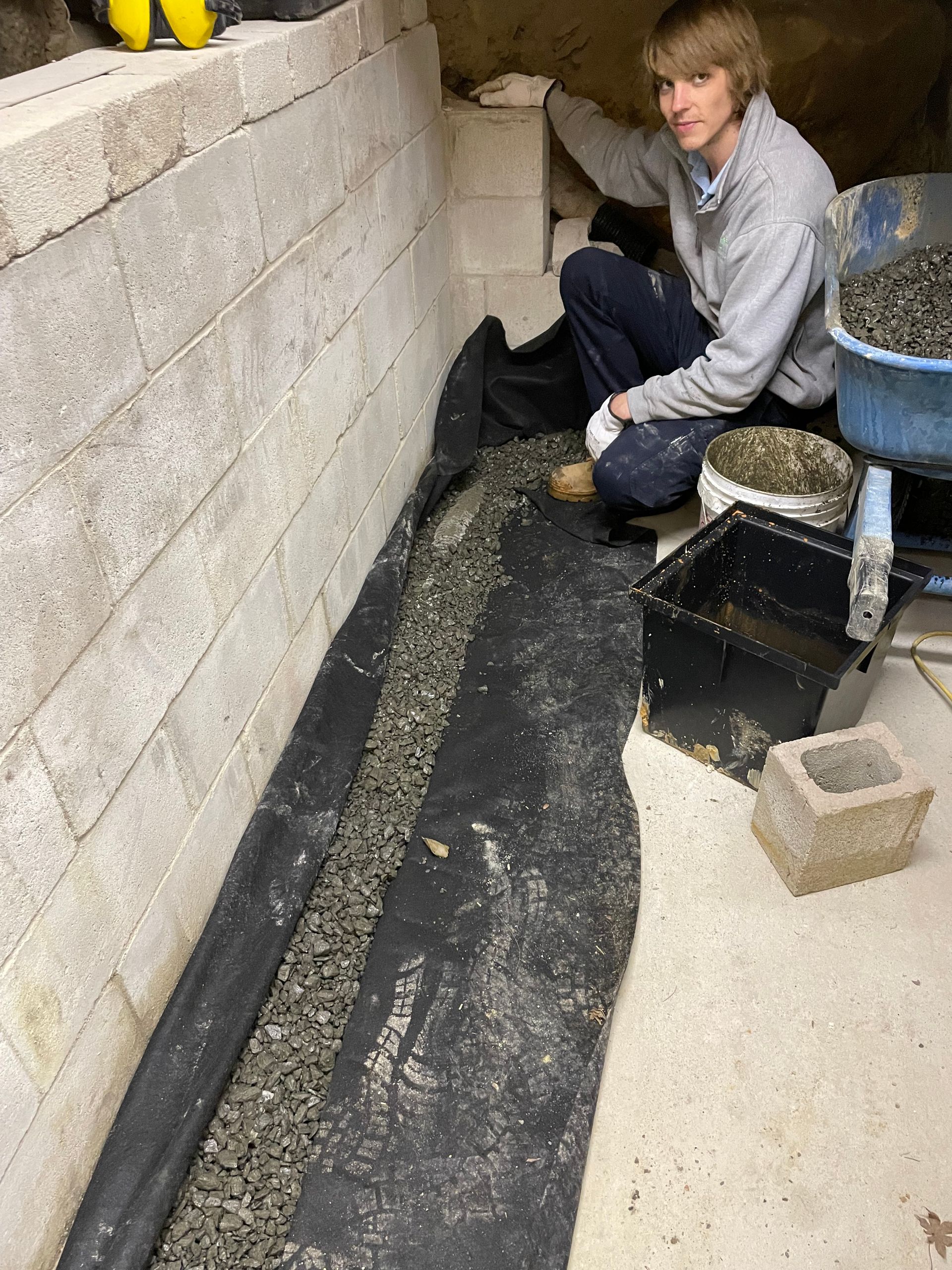 Man kneeling, installing gravel and black fabric in a basement next to concrete block wall.