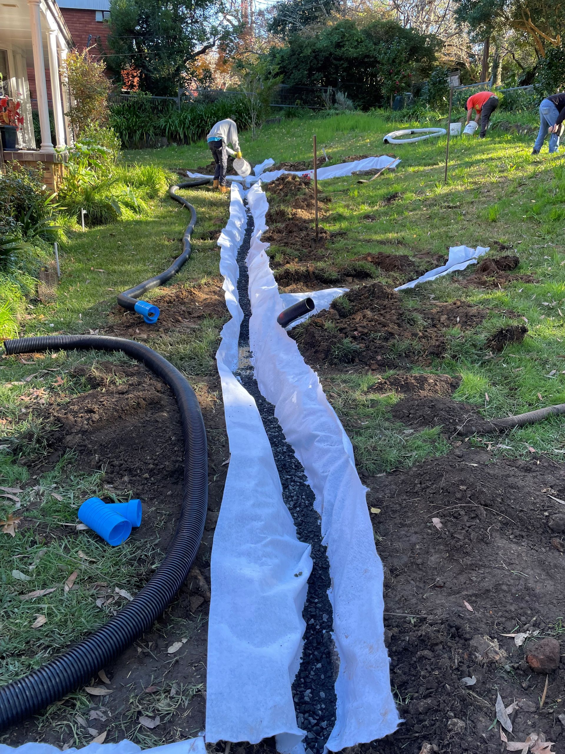 Workers install a drainage system in a grassy yard. Black pipe and gravel are laid within a white fabric-lined trench.