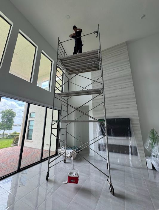 Worker on tall scaffolding inside a bright room, painting or repairing the ceiling.
