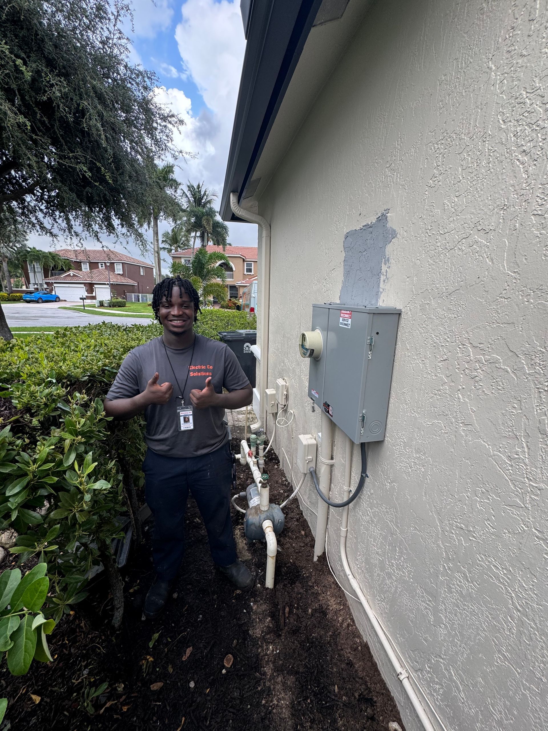 Person standing beside a house wall with utility meters and pipes, holding a tool outdoors.