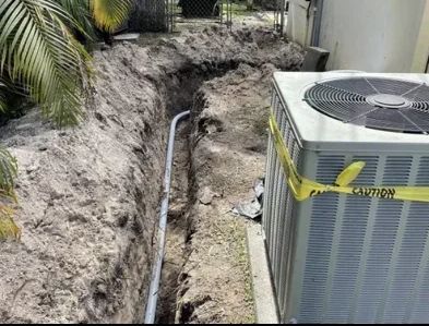 Trench dug beside an air conditioner unit, exposing buried pipes in a sandy yard.
