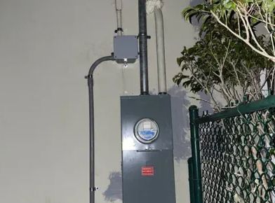 Gray electrical meter box and conduit mounted on a wall beside a fence and tree.