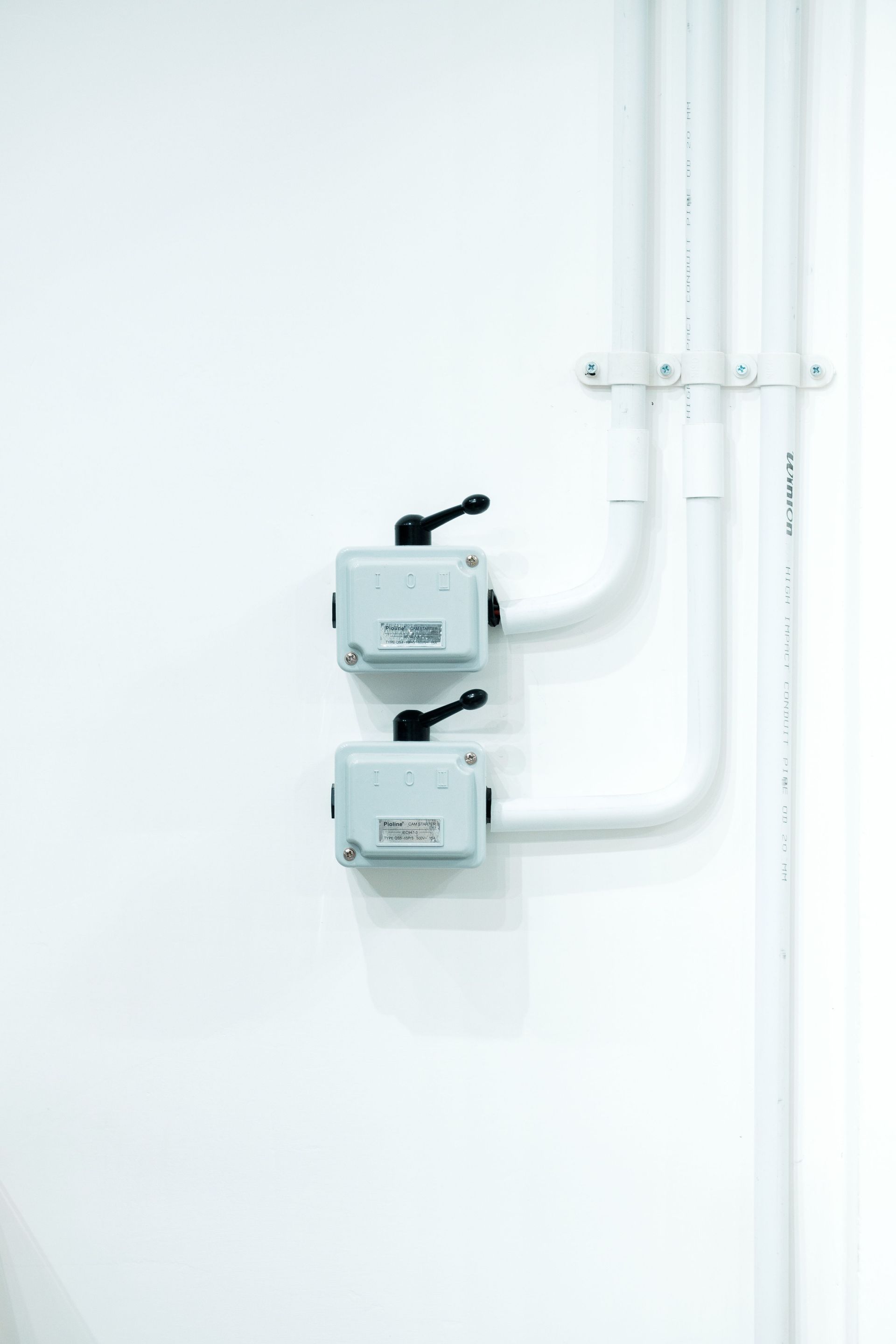 Two small gray wall-mounted electrical boxes with black switches and white conduit on a plain white wall