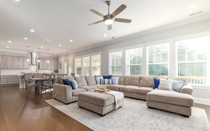 Bright open-concept living room with beige sectional, gray ottoman, and kitchen in the background