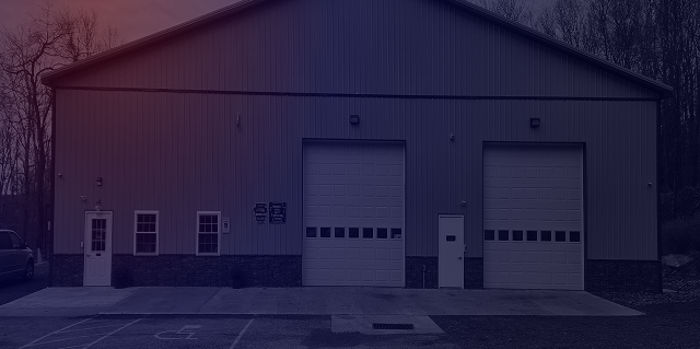 Small gray metal warehouse with two white garage doors and front windows, seen in dim light. | Hauger's Automotive Services