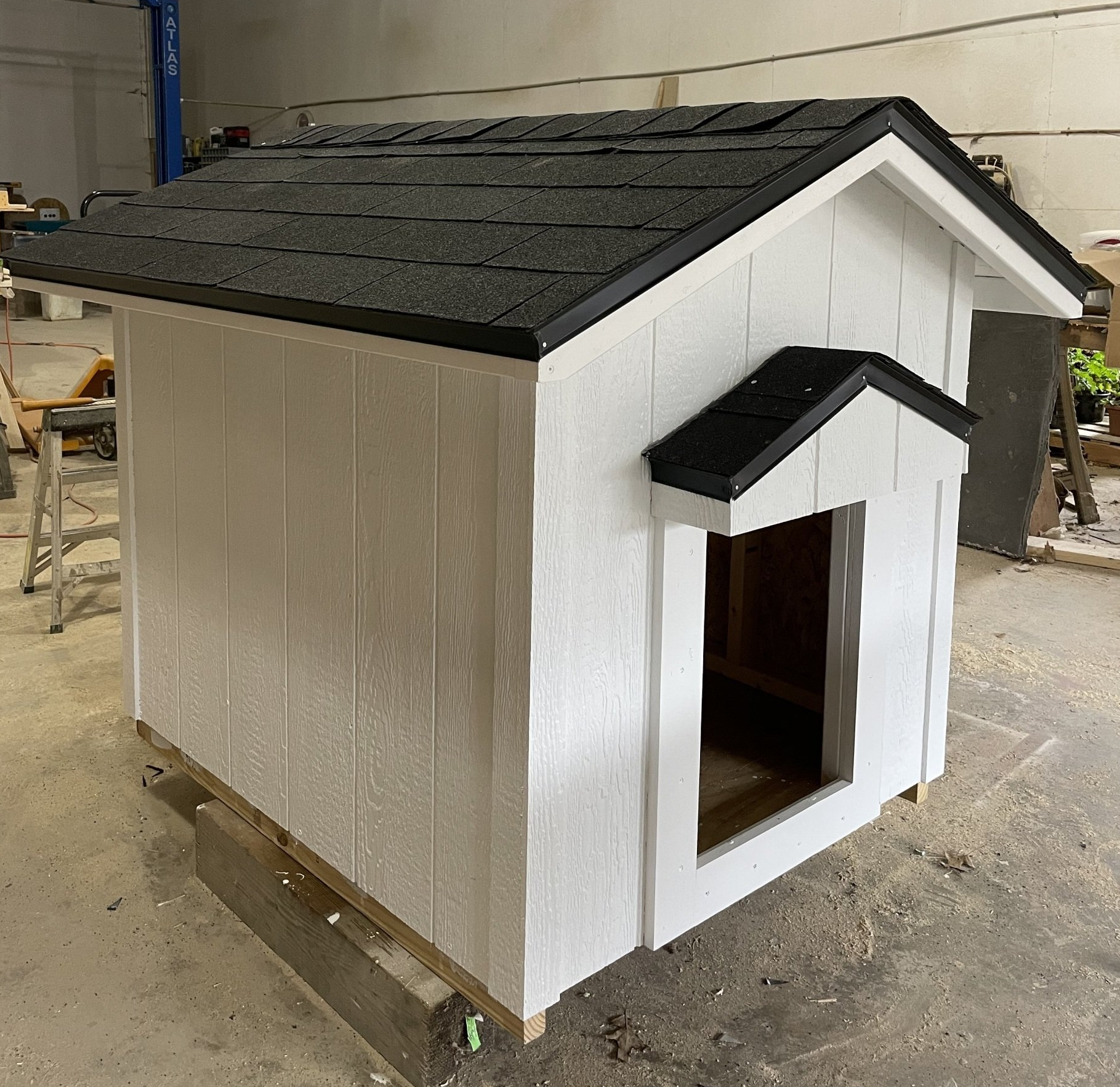 White dog house with black roof and trim, sits on wooden beams indoors.