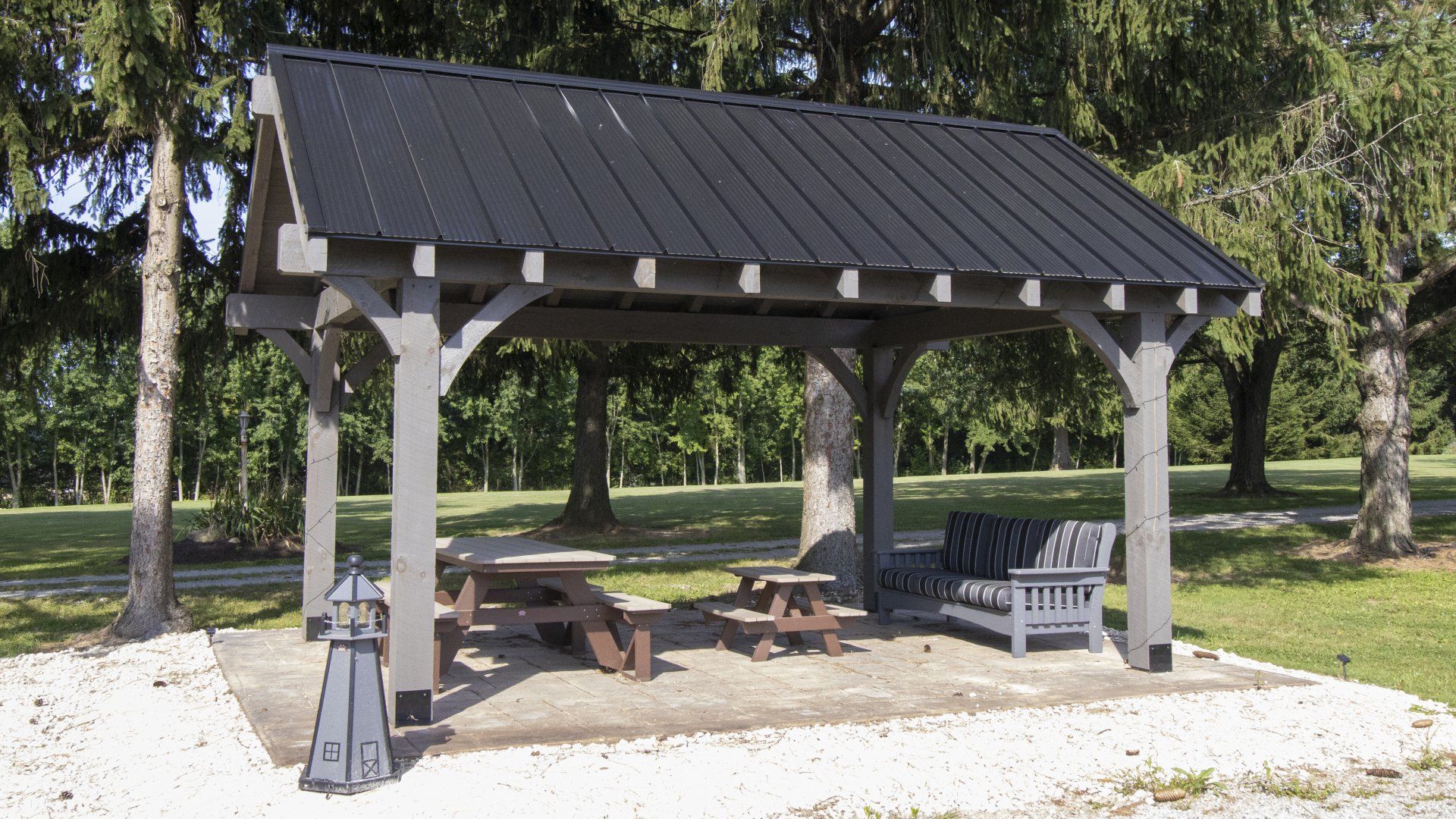 A gray wooden pavilion with a black metal roof, picnic tables, and seating in a park-like setting.