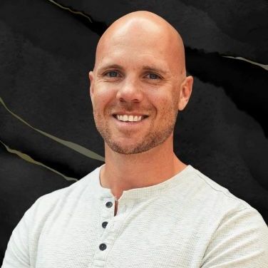 A bald man with a beard is wearing a white shirt and smiling.