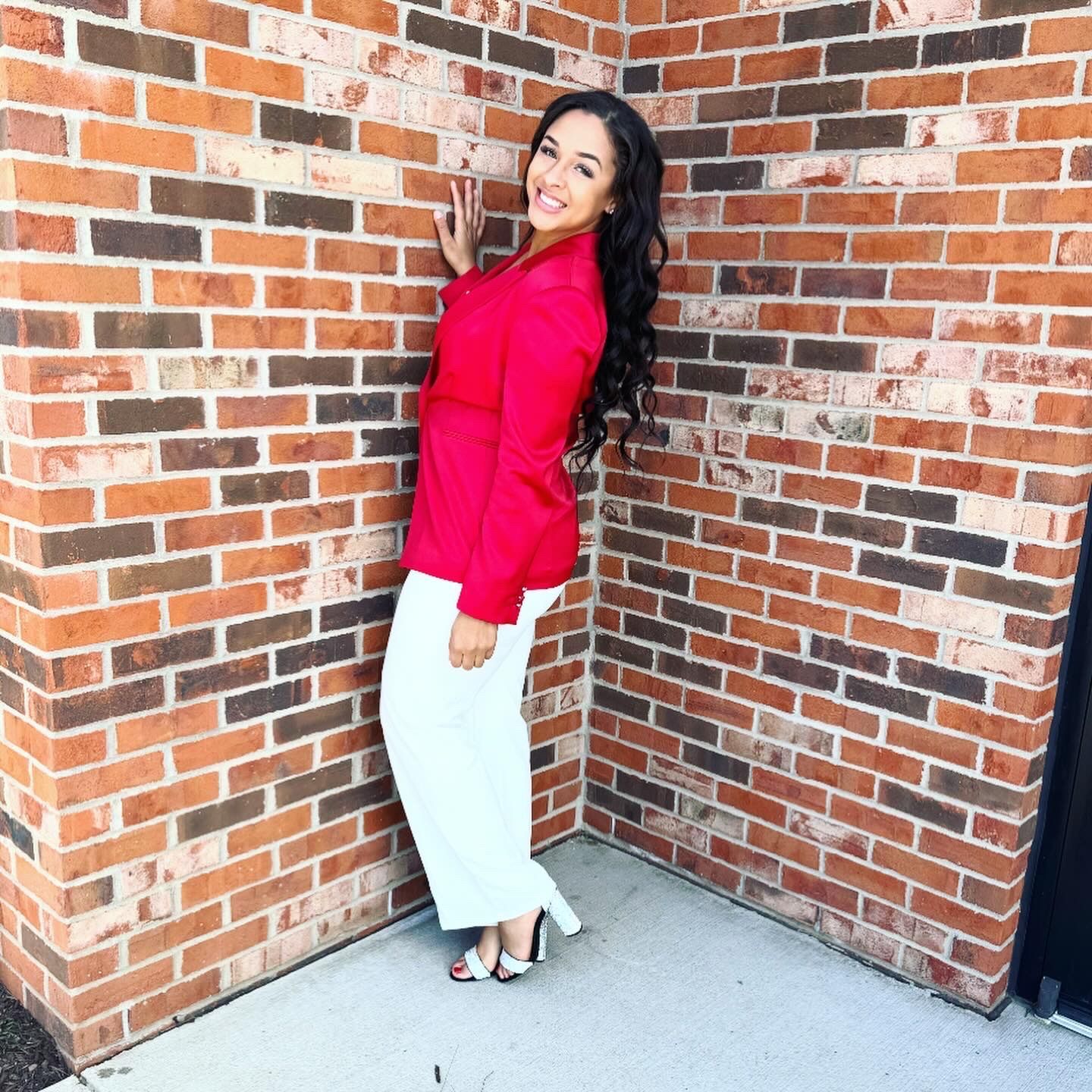 A woman in a red top and white pants is standing in front of a brick wall.