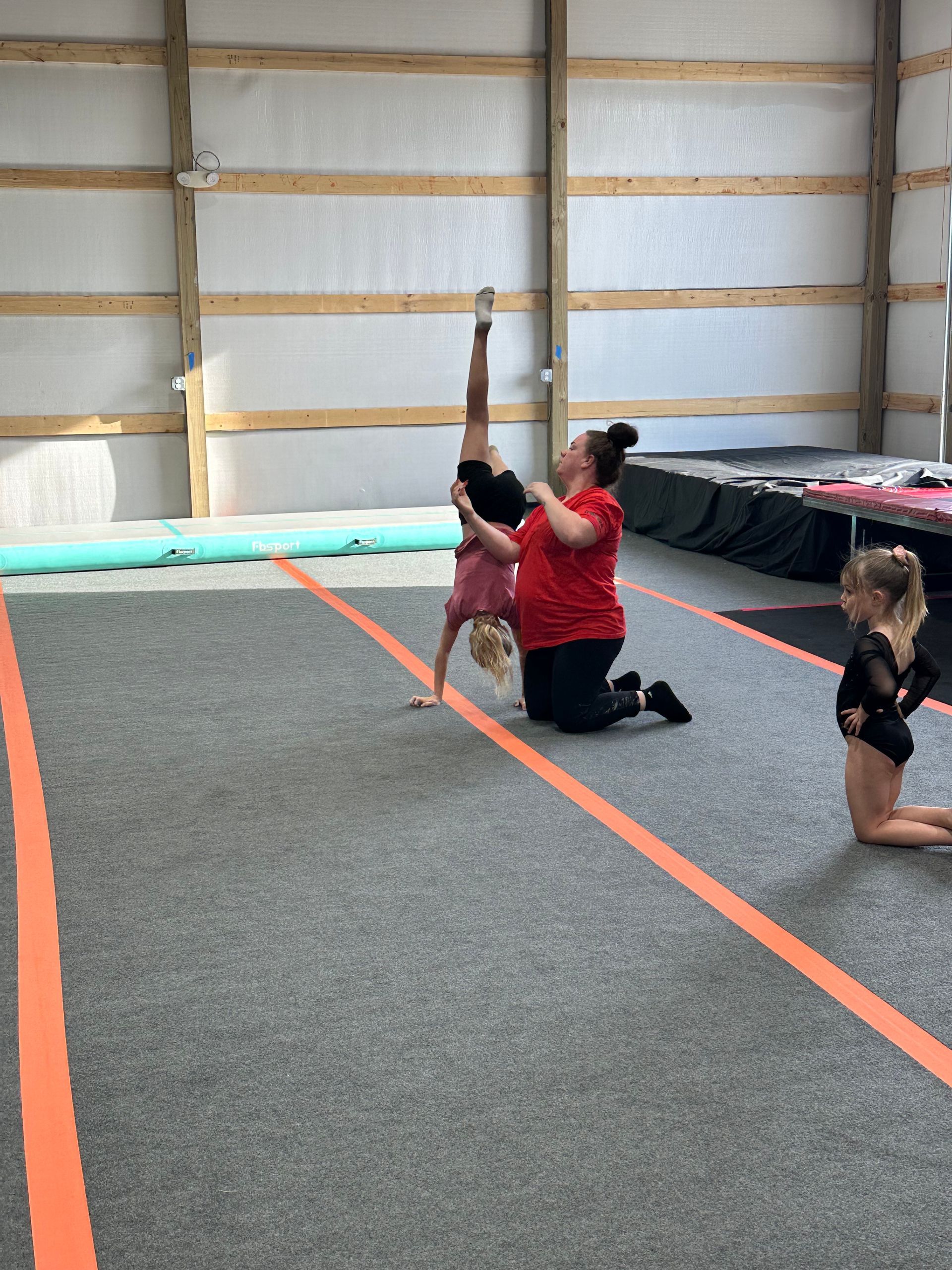 Gymnast doing a handstand, assisted by a coach, on a mat; another child kneels nearby.