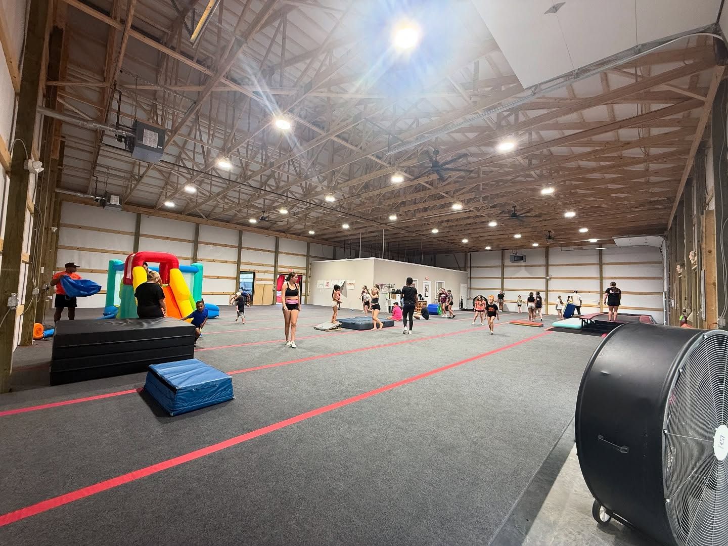 Large indoor gym with people training; includes mats, inflatable obstacle, and large fan.