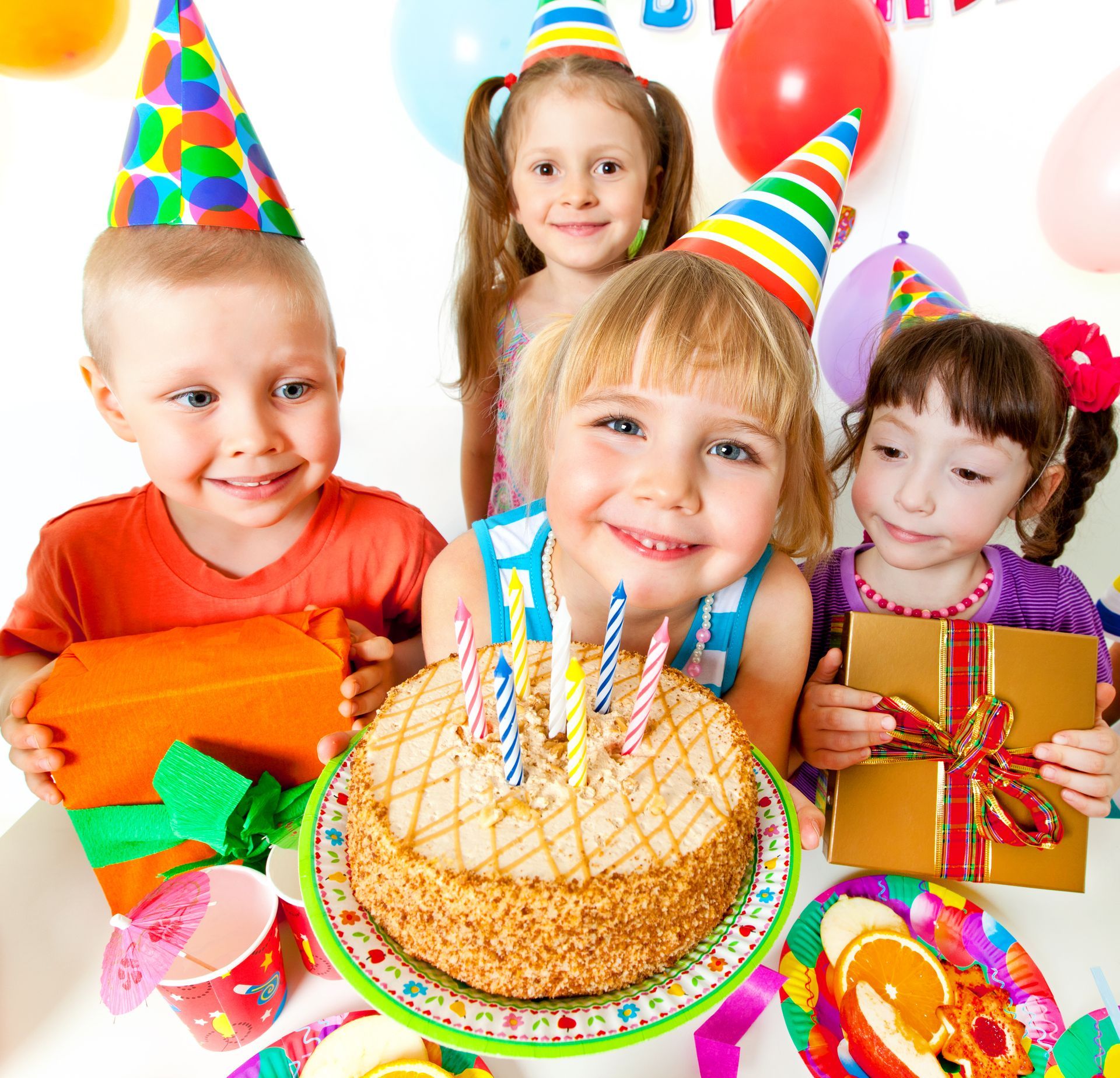 Children at a birthday party smiling with cake, presents, balloons, and party hats.