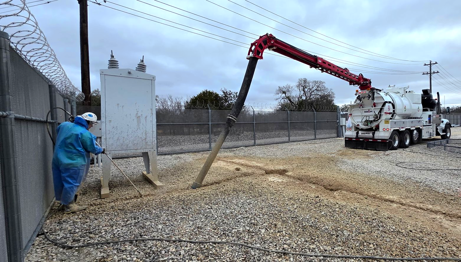 Worker in blue suit near fence as red boom truck pours gravel into a gravel yard