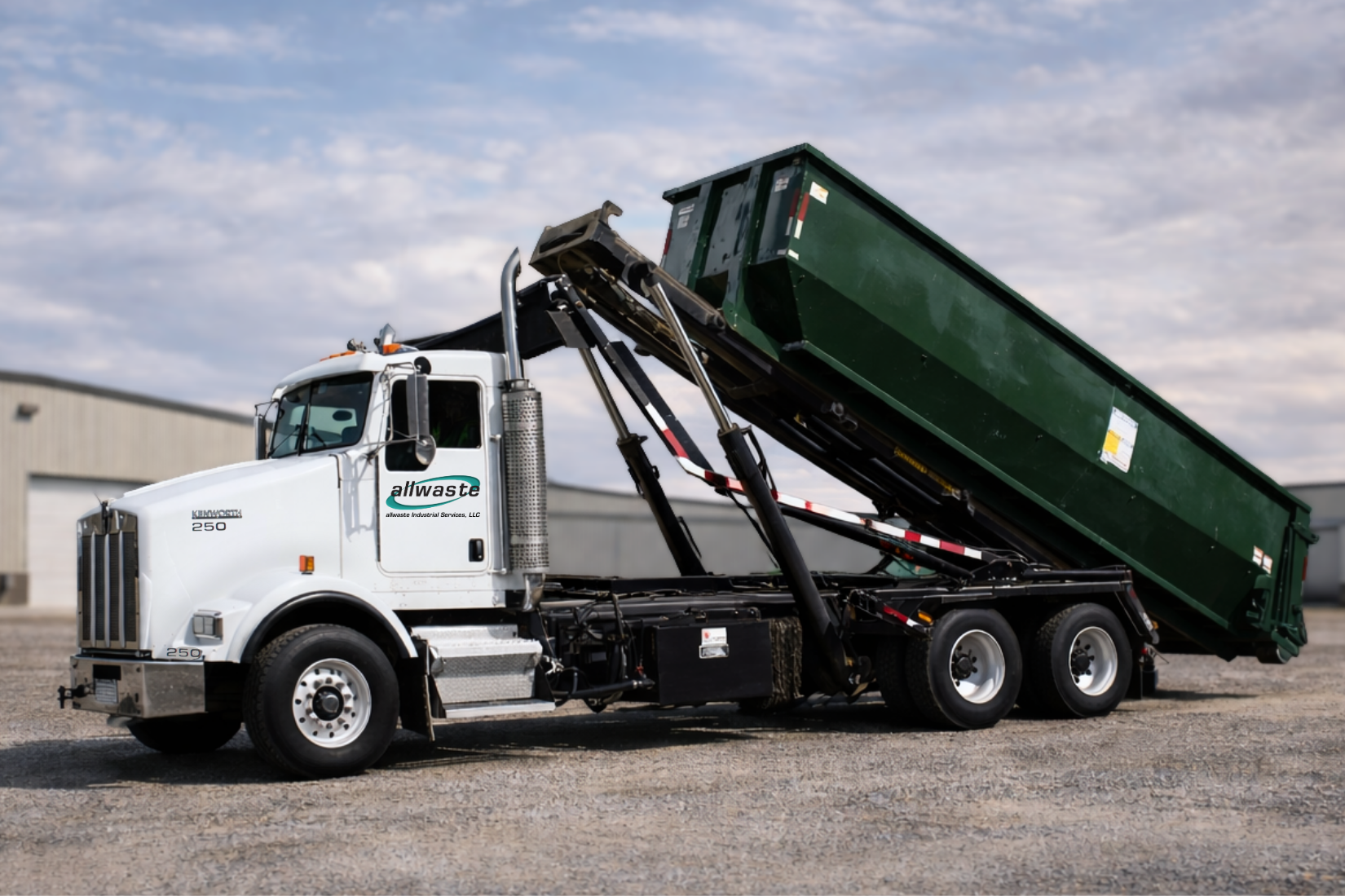 A white Kenworth roll-off truck in an industrial lot with its large green container tilted in the process of unloading.