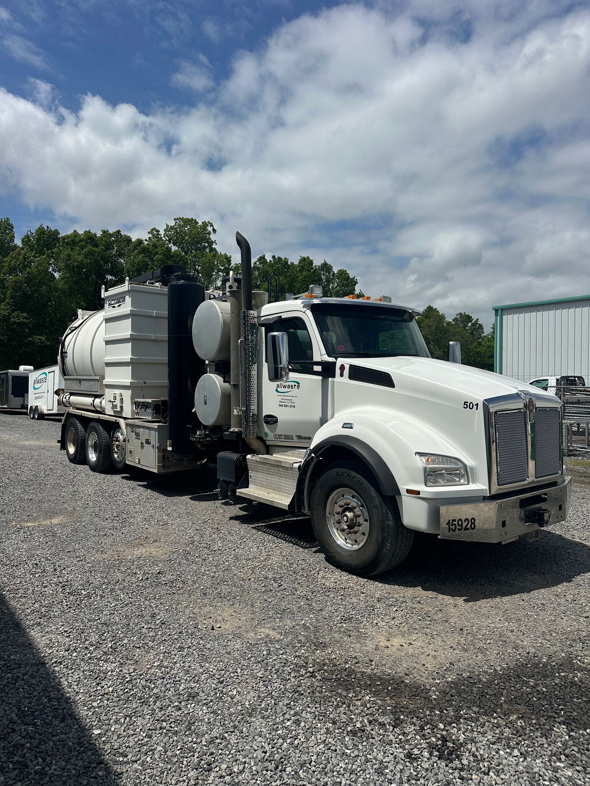 A white vacuum truck parked on a gravel lot near industrial equipment under a cloudy sky.