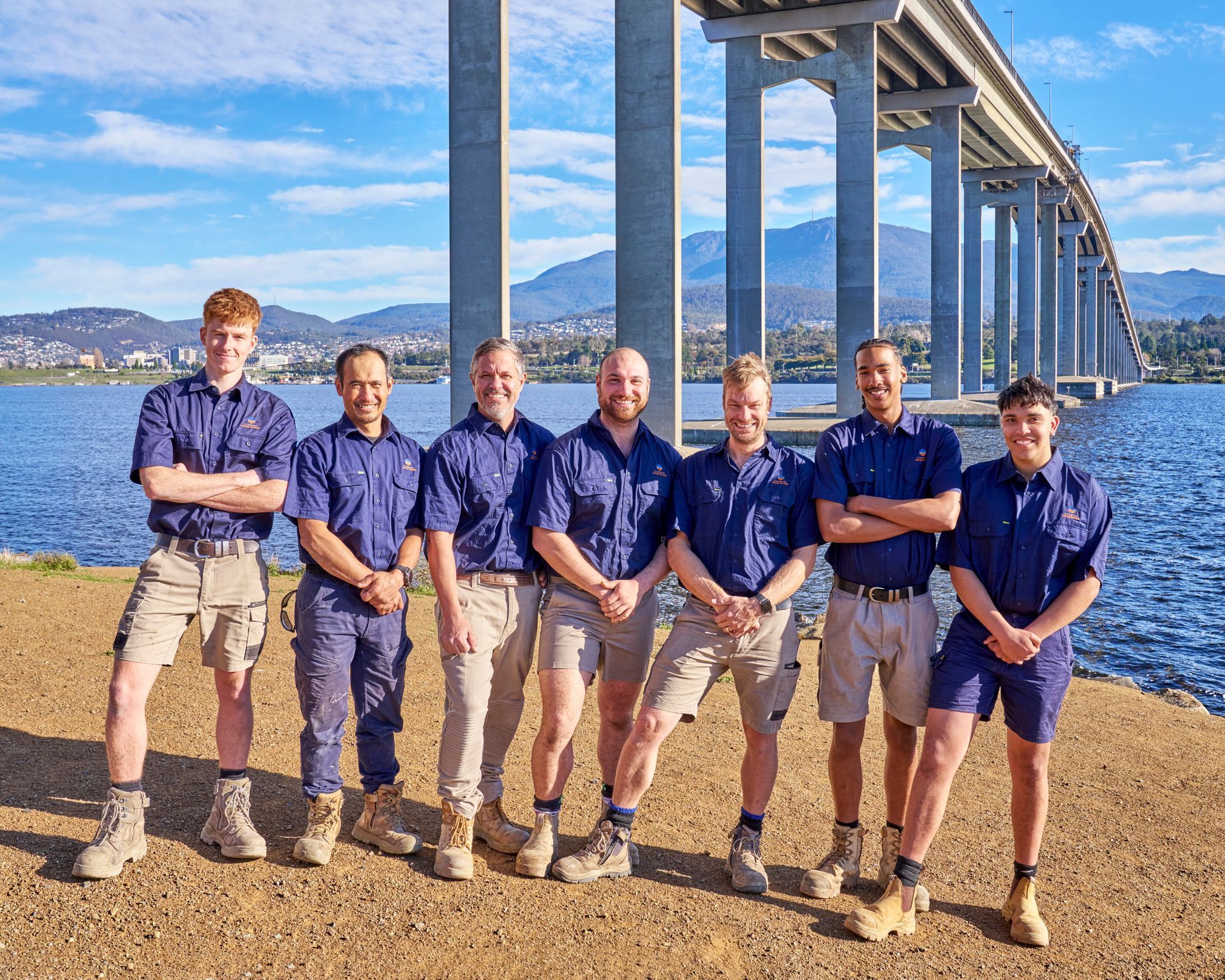Six men in matching blue shirts pose by a waterfront with mountains and a bridge behind them — Confac Energy Services In Lenah Valley, TAS