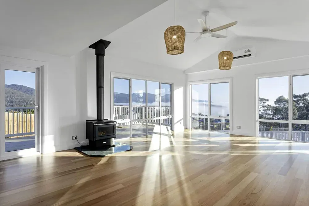 Bright modern living room with wood floors, white walls, large windows, and a black wood stove.— Confac Energy Services in Moonah, TAS
