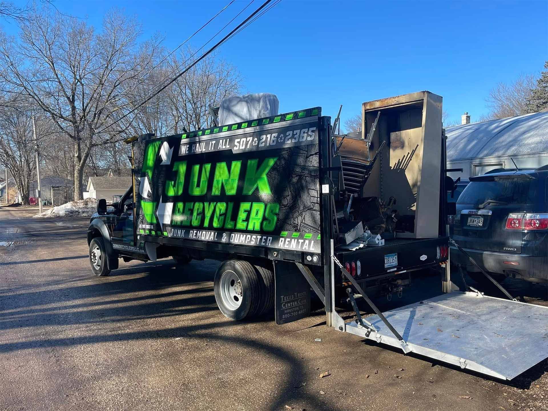 Junk removal truck with loading ramp, parked on a street, filled with debris. Green and black logo.