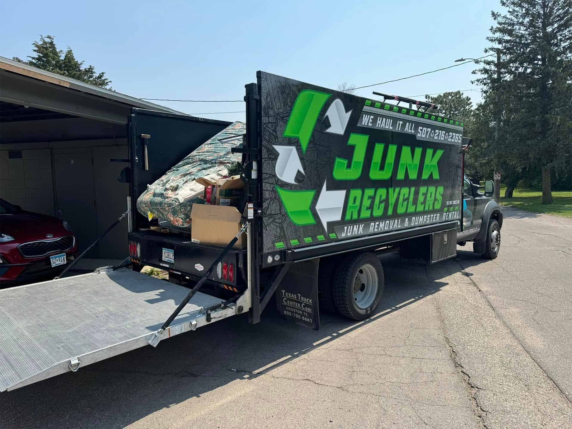 Junk recycler truck with open ramp and recycling logo, parked in front of garage on a sunny day.