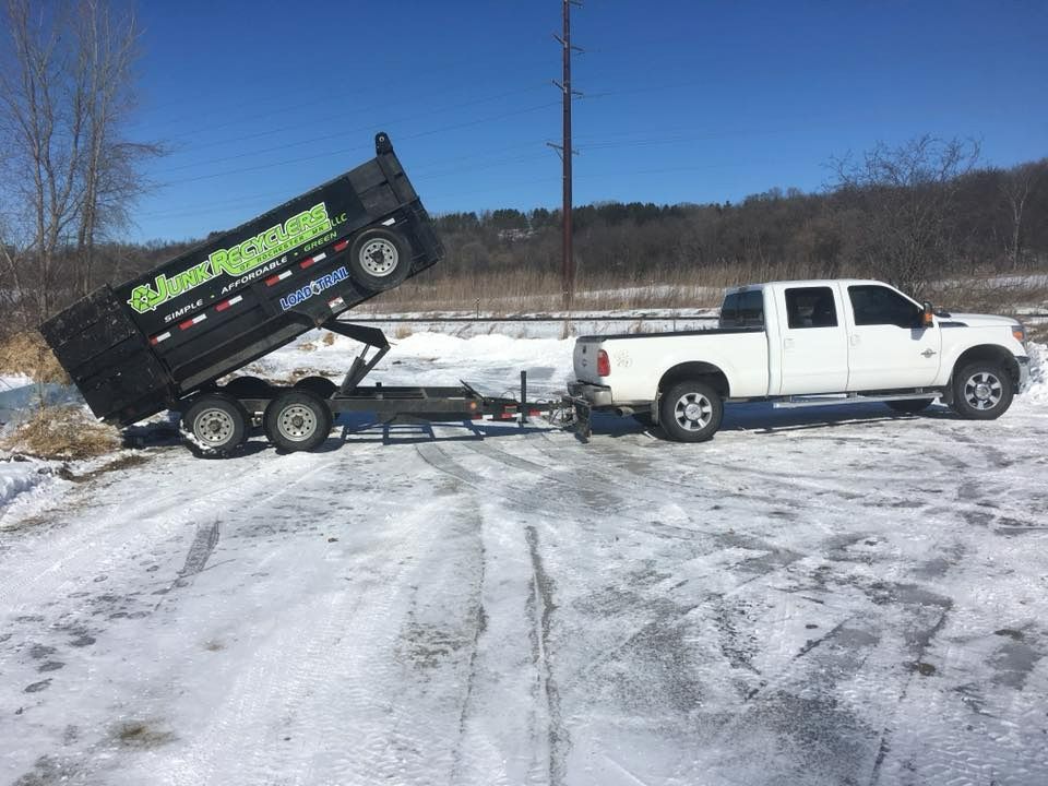 White truck towing a black dump trailer with the bed raised on a snow-covered dirt road