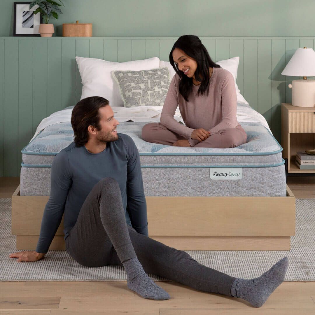 A person sits on the floor smiling at another person sitting on a mattress in a bedroom with green walls.